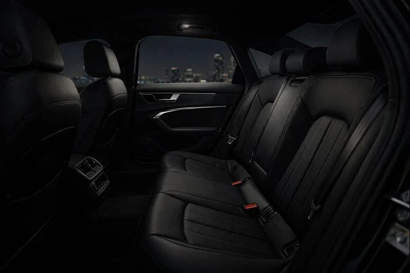 The interior of a car at night showing black leather back seats, side door with handle, window, and city skyline outside.