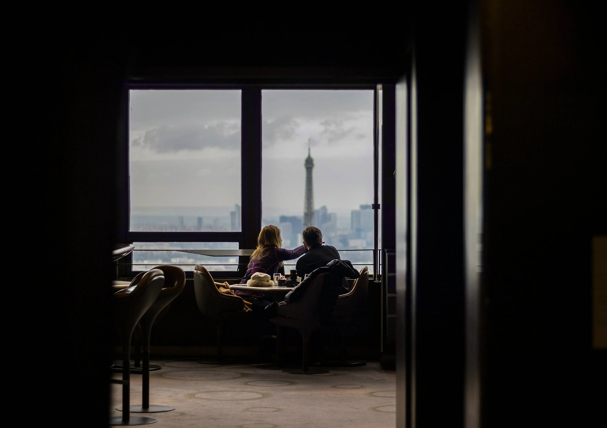View of two people sitting at a table in a high-rise cafe, looking out at the Eiffel Tower through large windows.