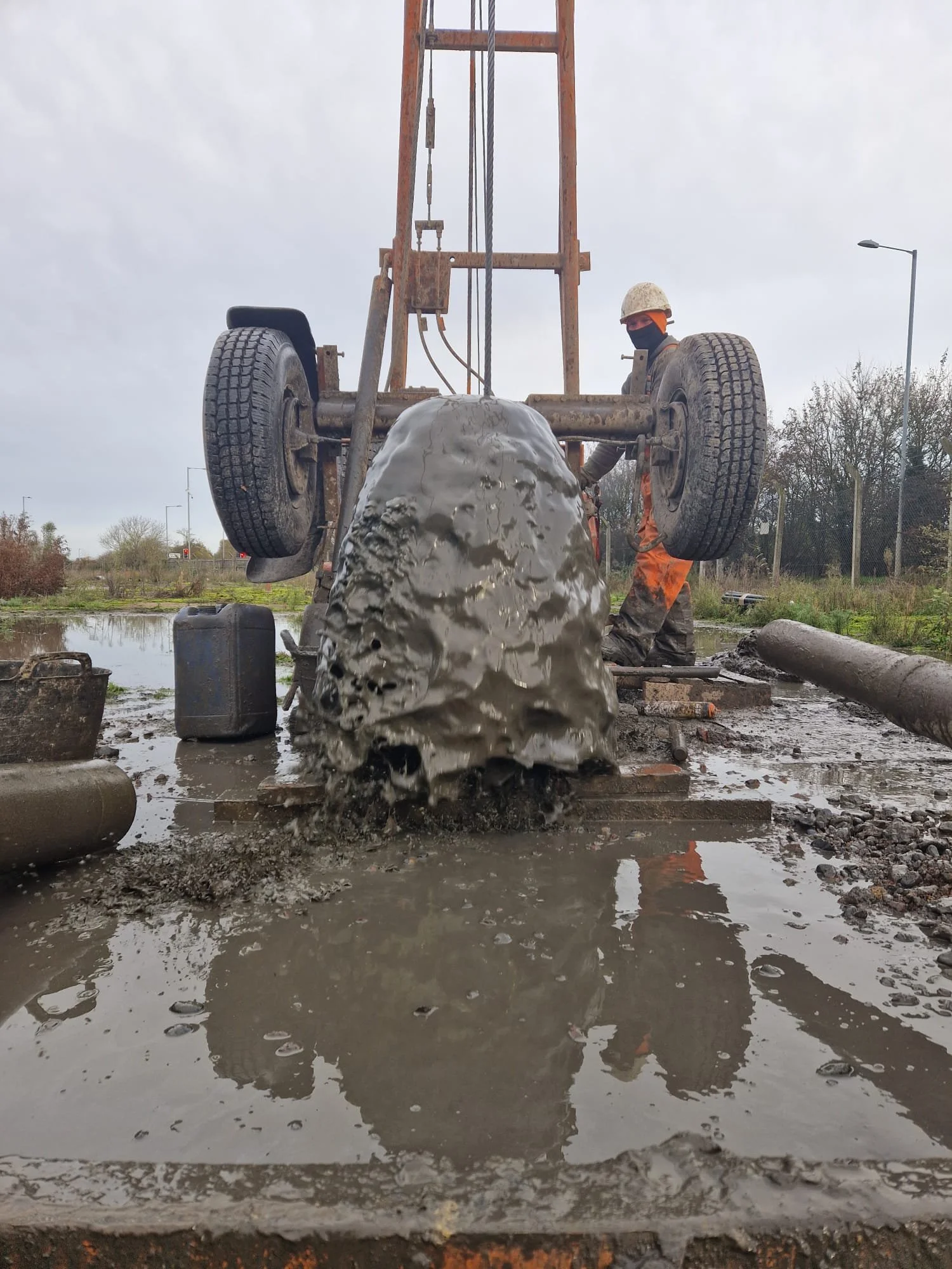 A worker in orange and black gear and a helmet is operating a mud drilling machine outdoors on a cloudy day. Mud is splashing as the machine drills into the ground.