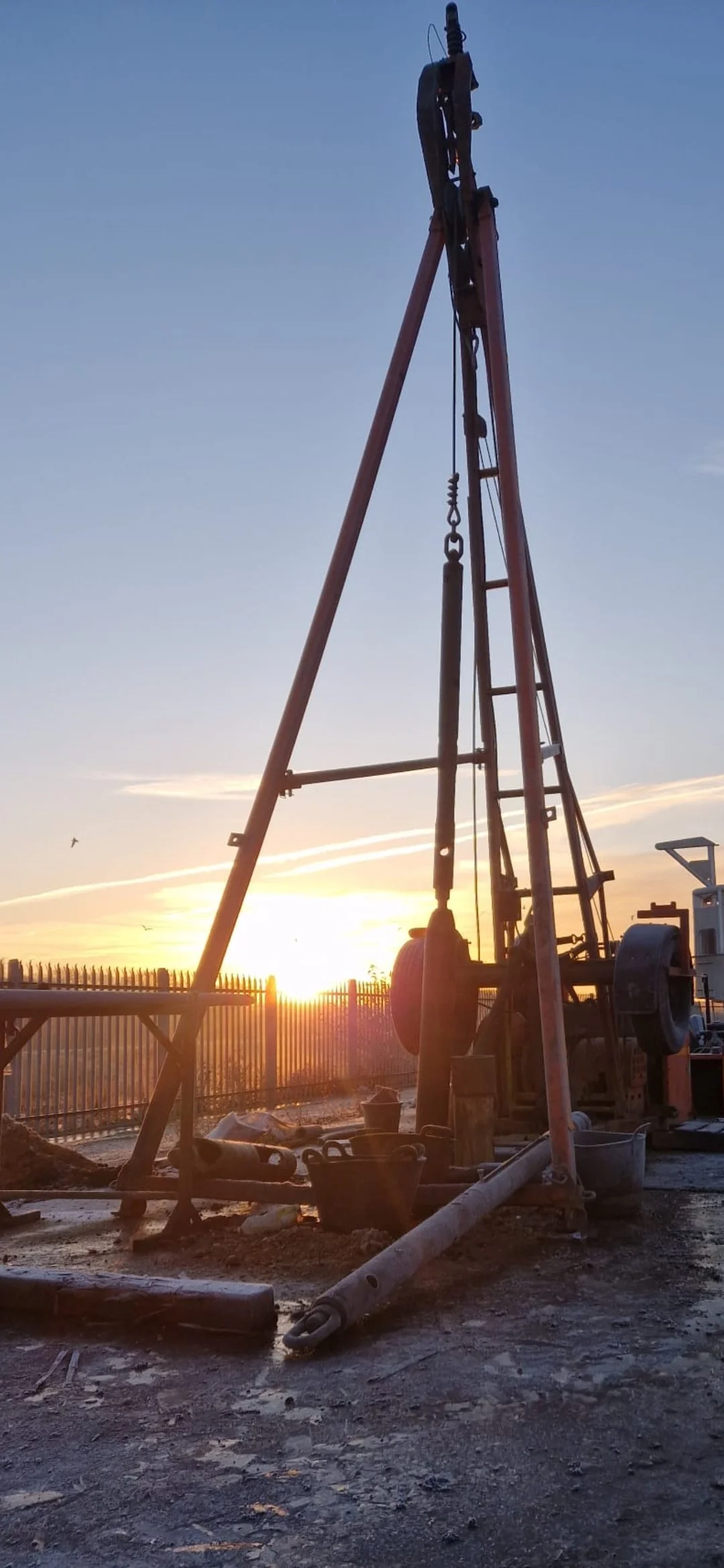 A drilling rig silhouetted against a sunset sky, situated on a construction site with tools and barrels around.