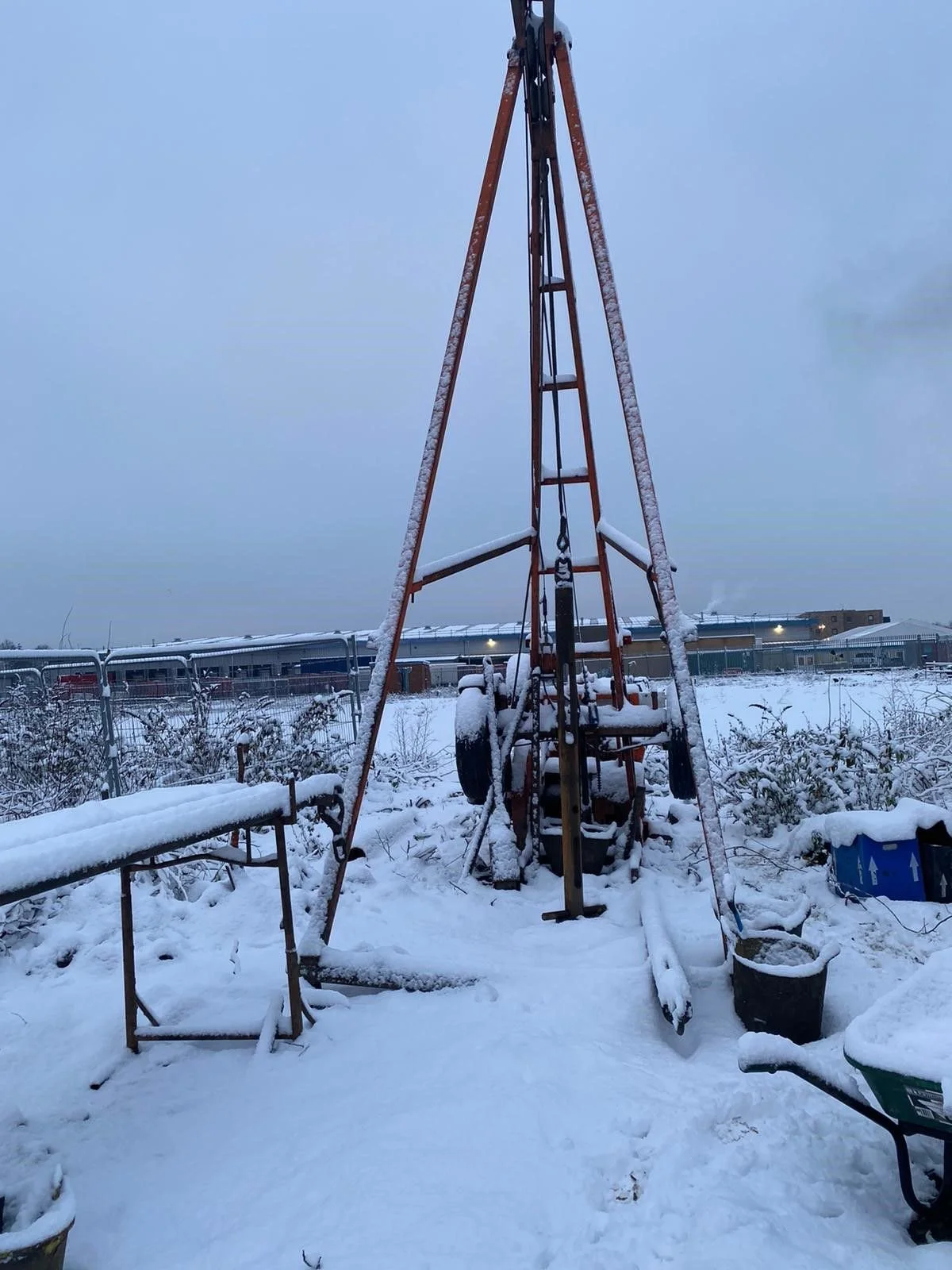 Snow-covered outdoor construction or drilling equipment with a triangular metal frame, shovels, wheelbarrows, and buckets around it, in a snowy landscape.