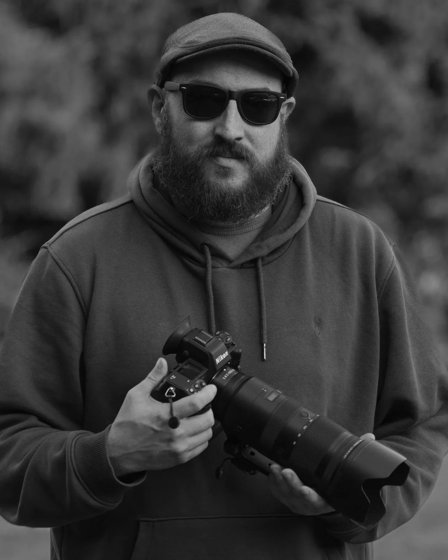 A man with a beard wearing sunglasses, a cap, and a hoodie holding a professional camera in an outdoor setting.