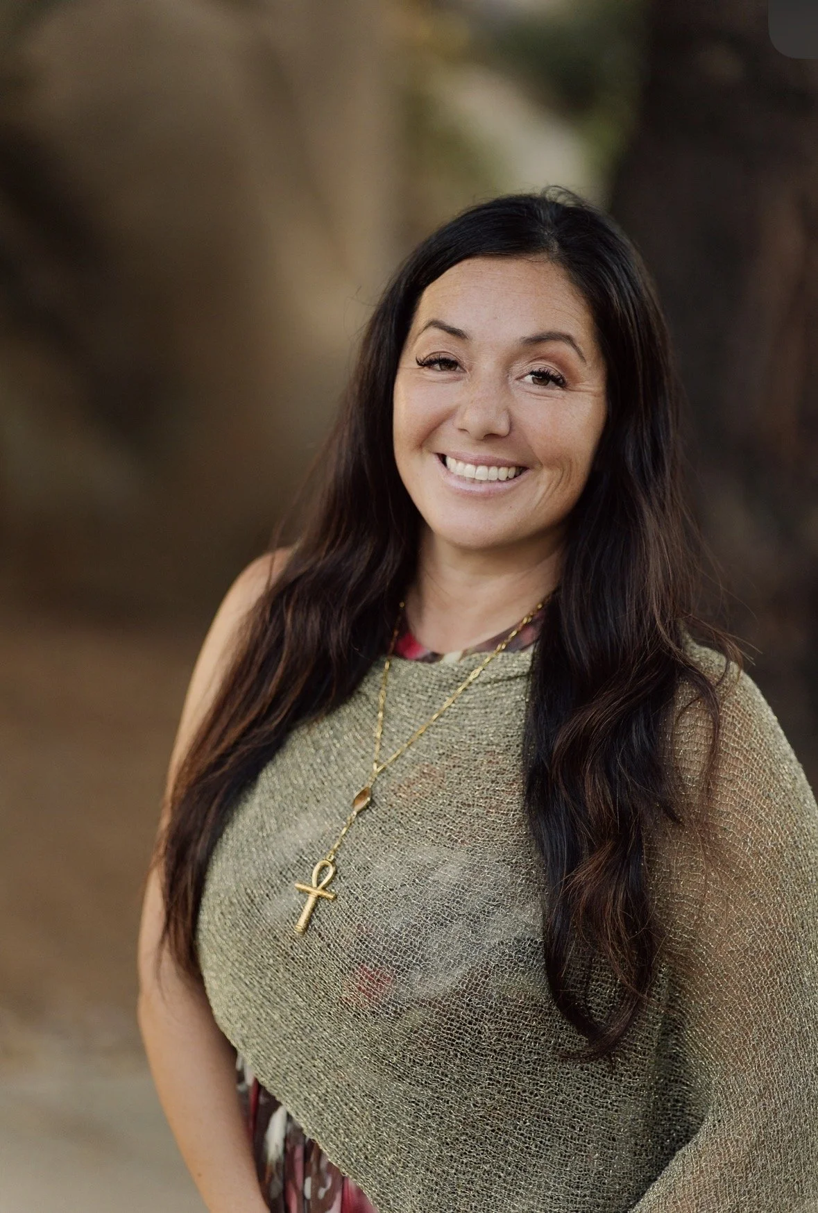 A woman with long dark hair smiling outdoors, wearing a gold necklace with an ankh pendant, a textured gold top, and a floral dress underneath.