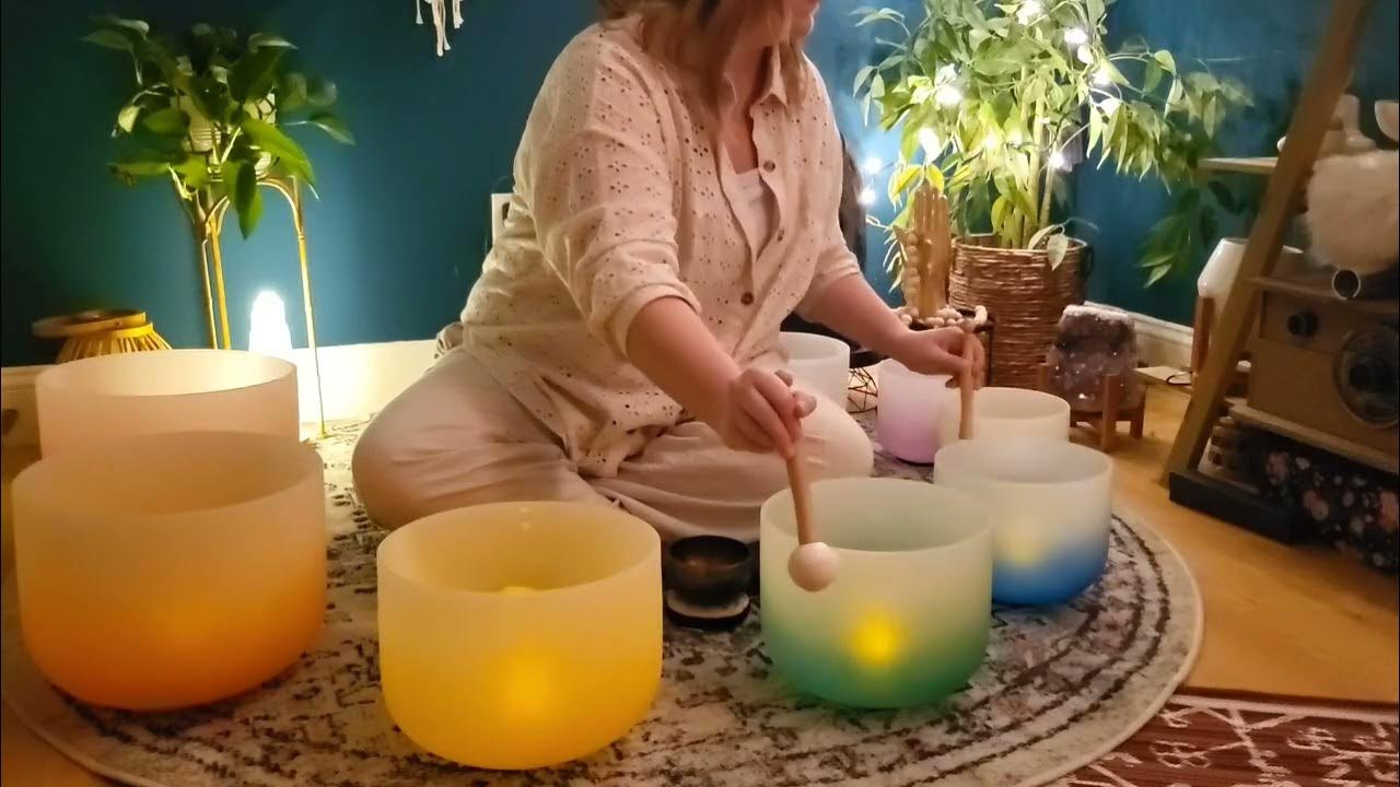 Person sitting on a patterned rug playing crystal singing bowls with mallets in a cozy room decorated with plants and soft lighting.