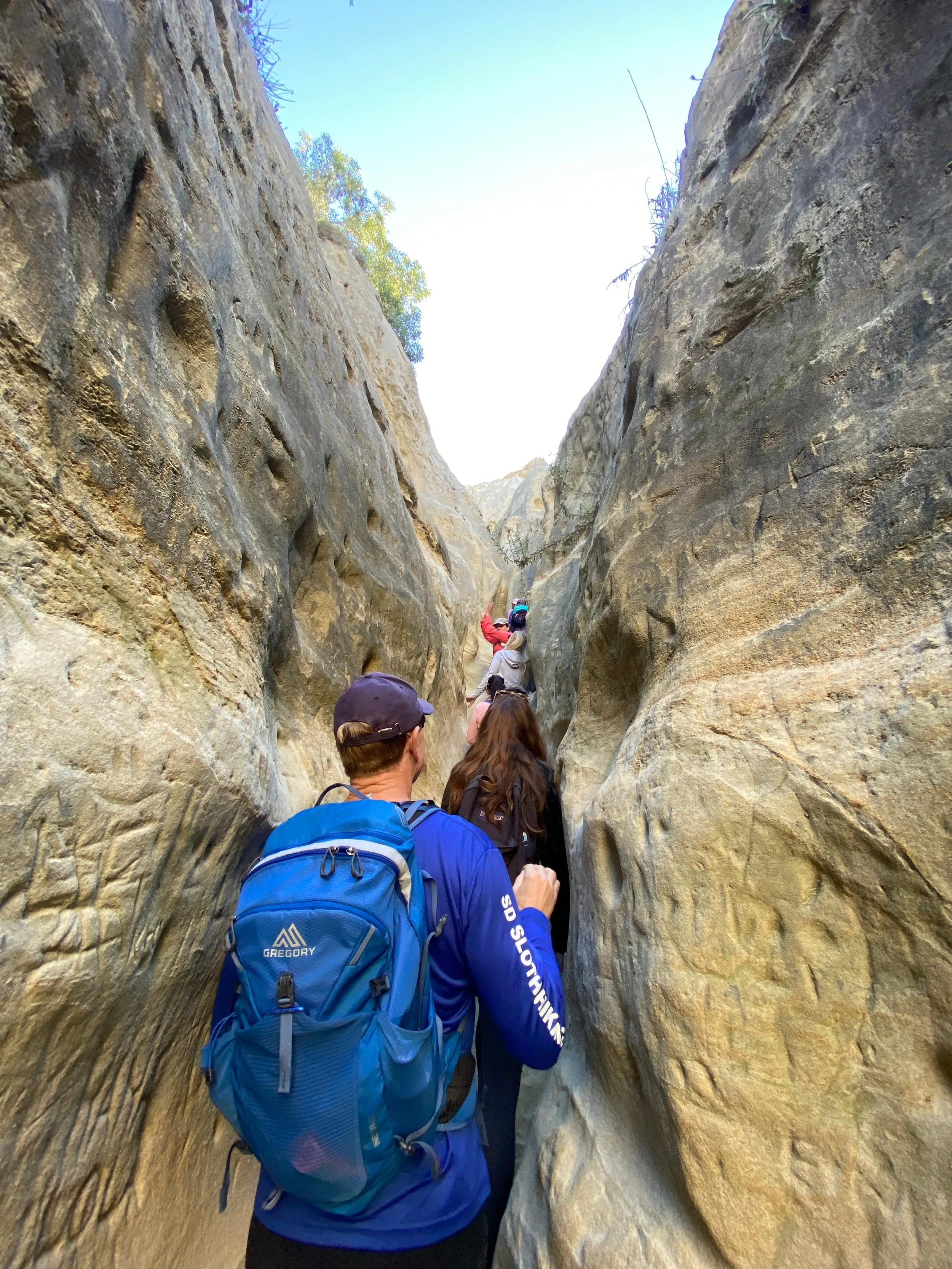 Group of hikers with backpacks walking through a narrow canyon with tall, smooth rock walls on either side.