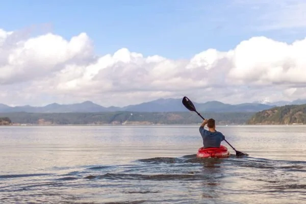 Person kayaking on a calm lake with mountains and clouds in the background.