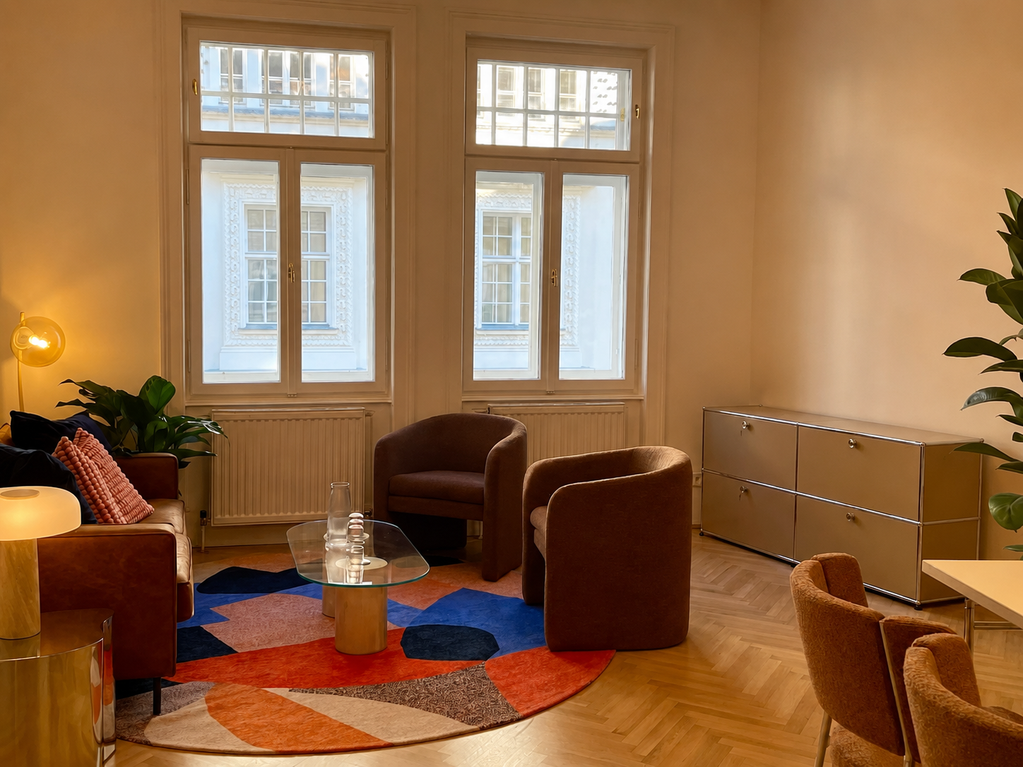 Einladender Therapieraum  mit großen Fenstern, braunen Sesseln, einem bunten Teppich, einem weißen Sideboard und Pflanzen.