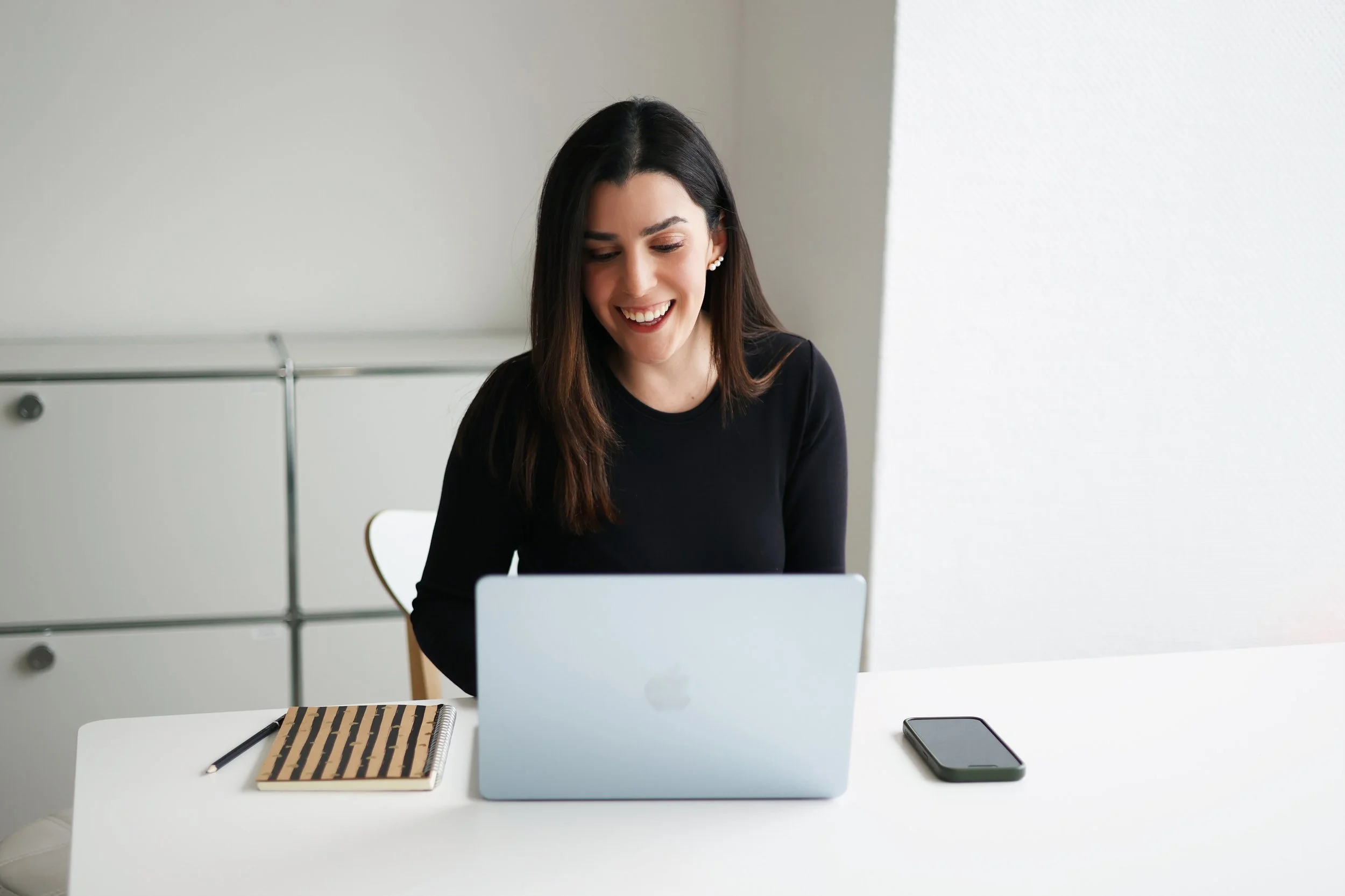 A young woman with dark hair smiling while working on a laptop at a white desk, with a notebook and pen on the left and a smartphone on the right.