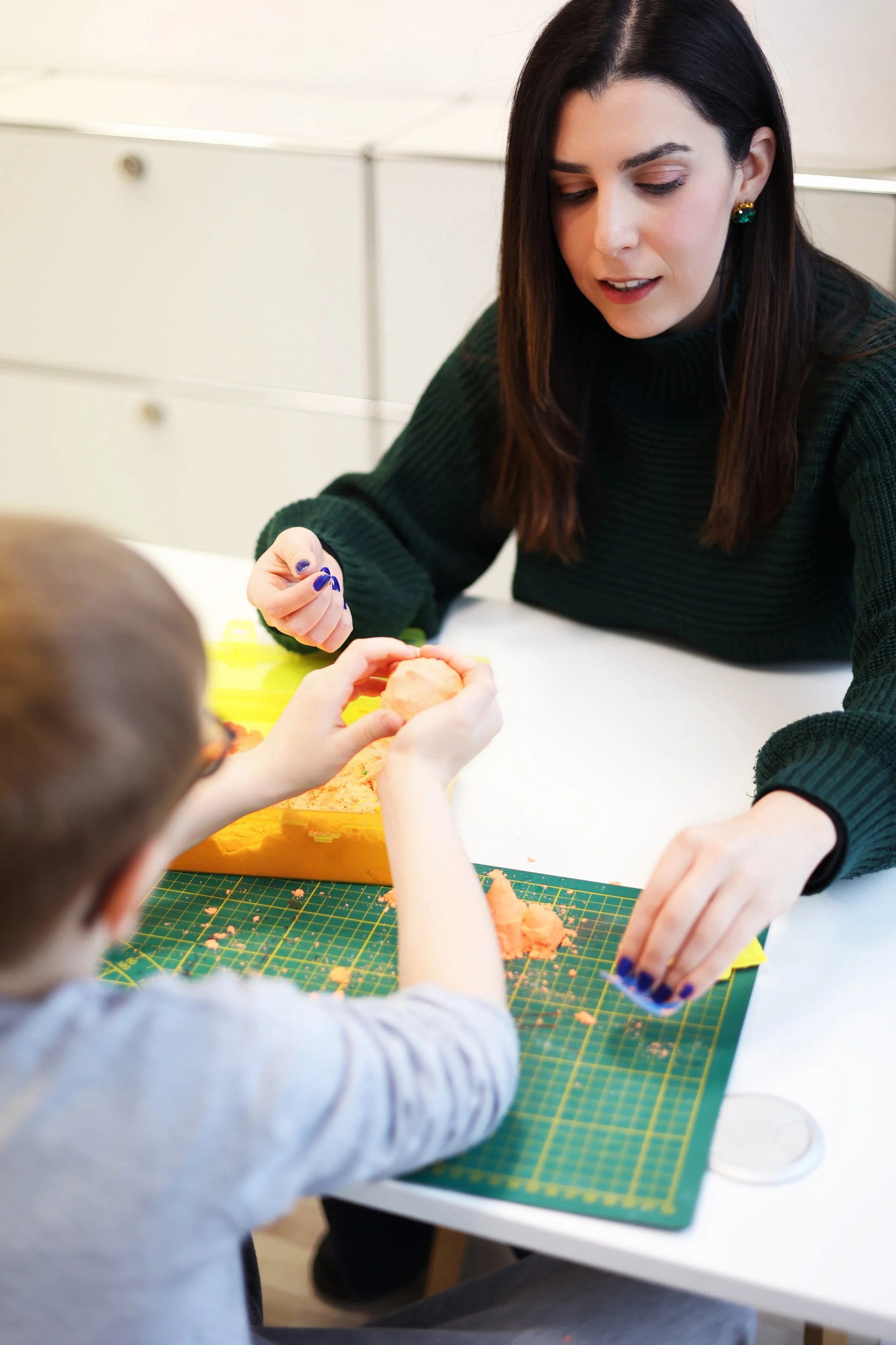 A woman and a child working together on a craft project at a table. The woman has dark hair, wears a green sweater, and is focused on helping the child. The child has short hair and is using their hands to shape orange clay or dough near a green cutting mat.