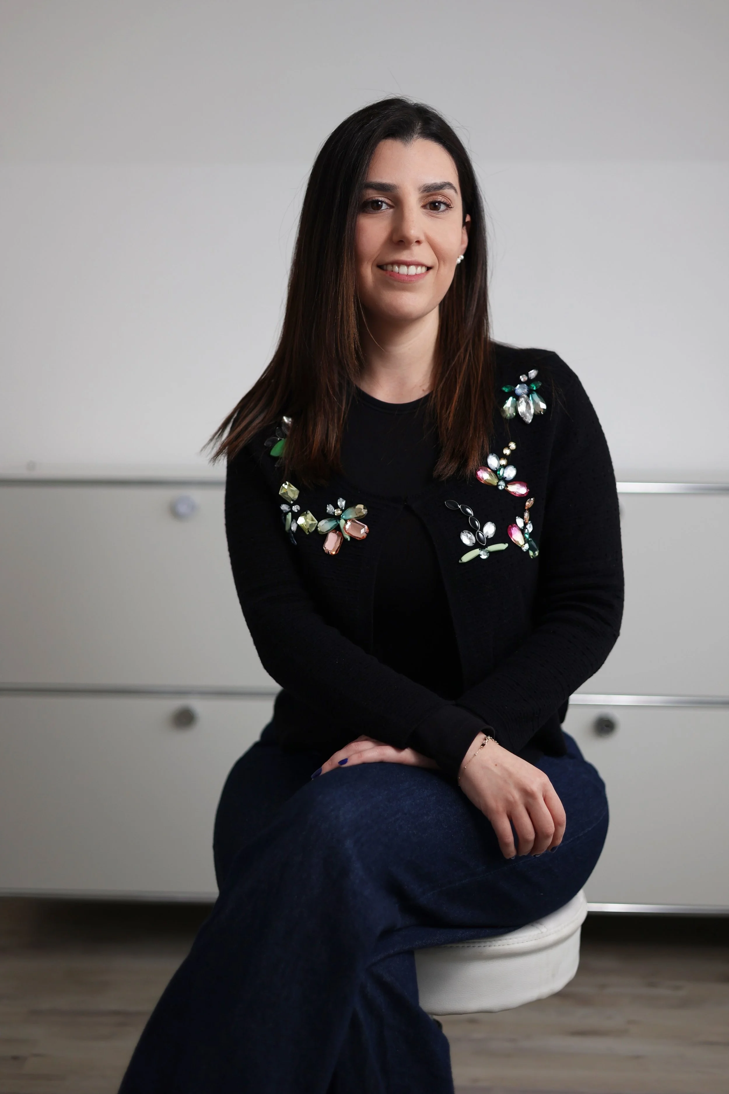 A woman with shoulder-length brown hair, wearing a black sweater with colorful embellishments, and navy blue pants, seated on a white chair in a room with a plain light-colored background.