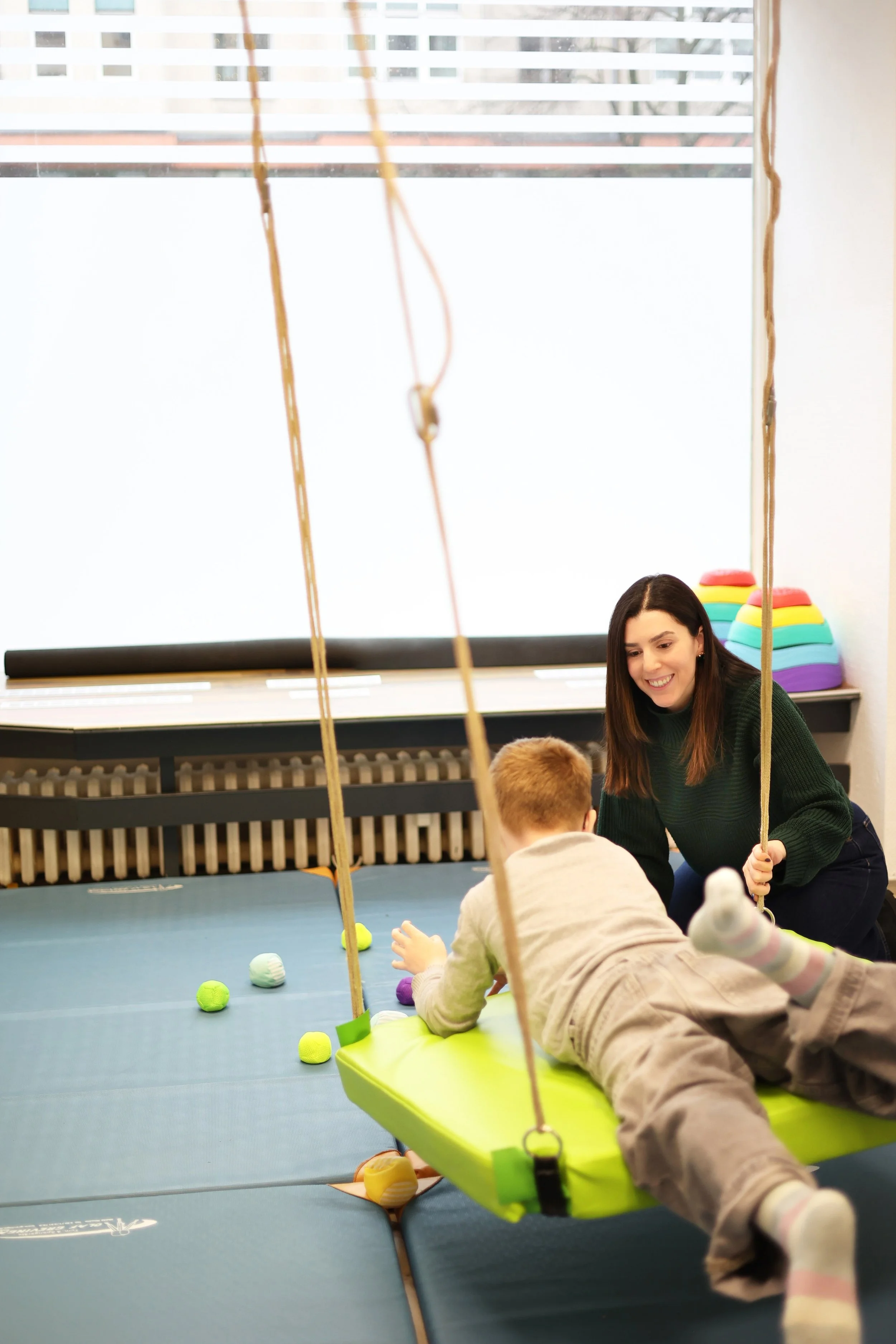 A woman smiling and supervising a young boy on a green swing indoors, with colorful foam shapes on the floor and a large window in the background.