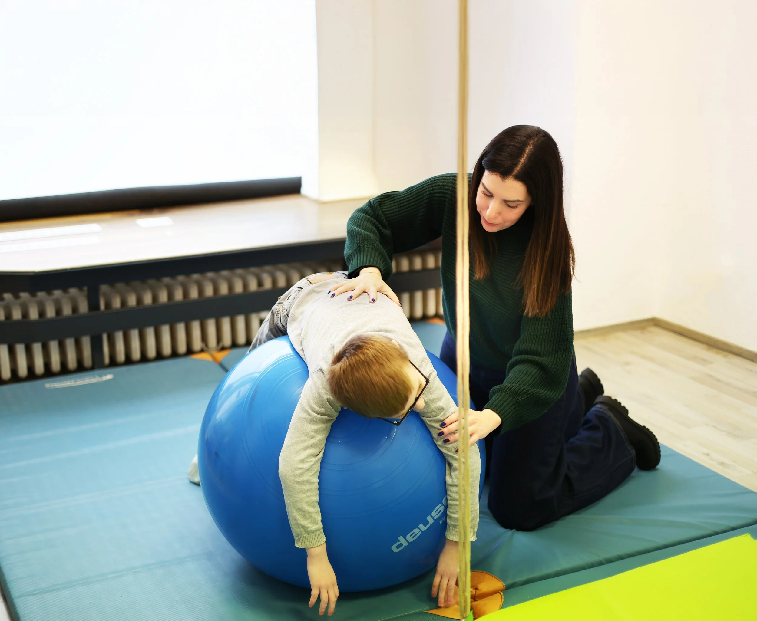 A woman assisting a child with balance on a large blue exercise ball during a physical therapy session in a clinical setting.