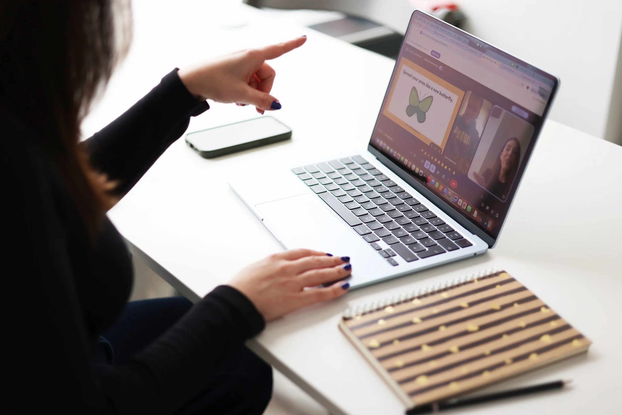 A woman sitting at a white desk, pointing at a laptop screen showing a video call, with a notebook and a smartphone on the desk.