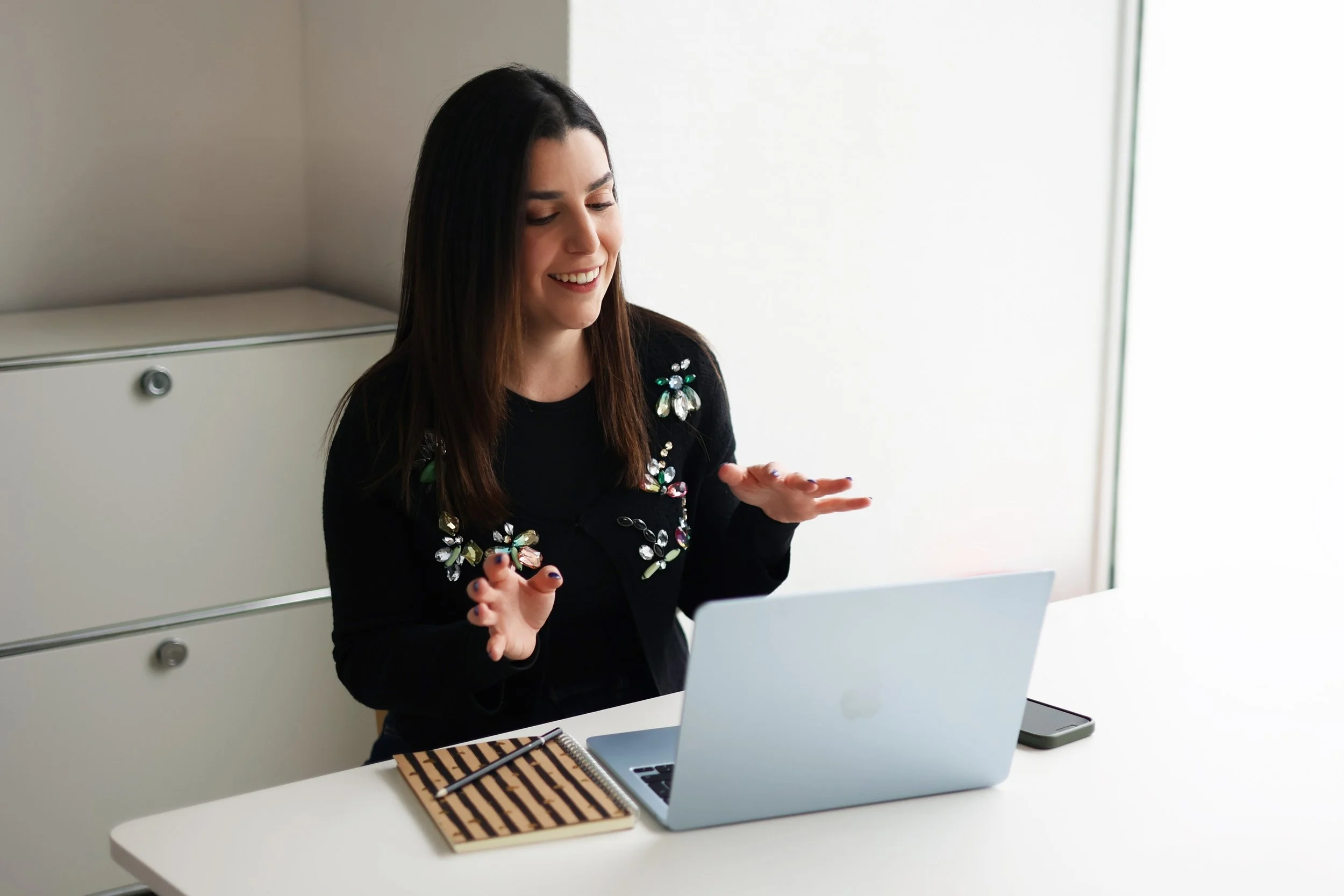 A woman smiling and gesturing with her hands while sitting at a white table with a silver laptop, a striped notebook, a pen, and a smartphone in front of her, in a bright, minimalist office.