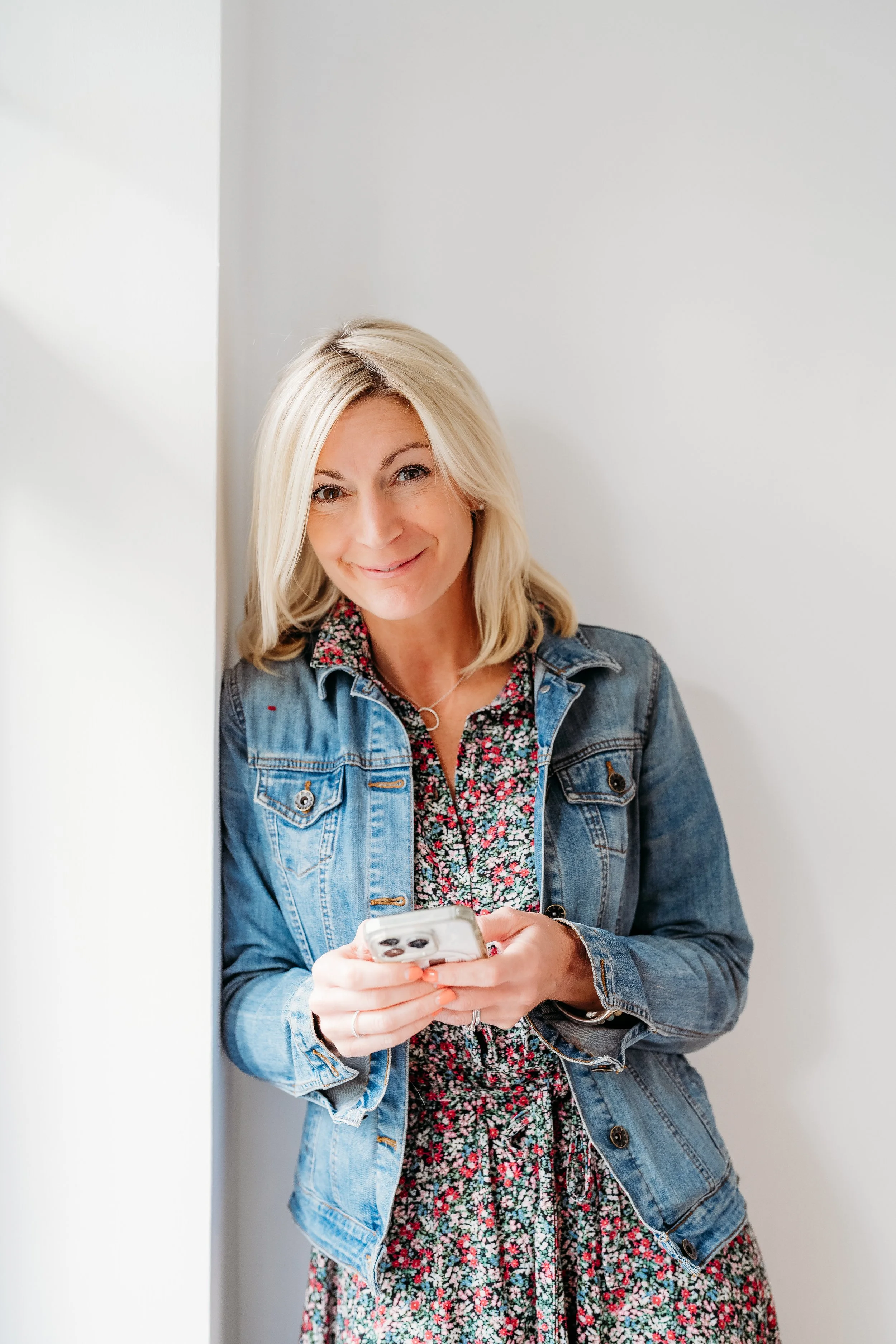 A woman with blonde hair smiling while looking at her phone, wearing a denim jacket over a floral dress, standing against a white wall.