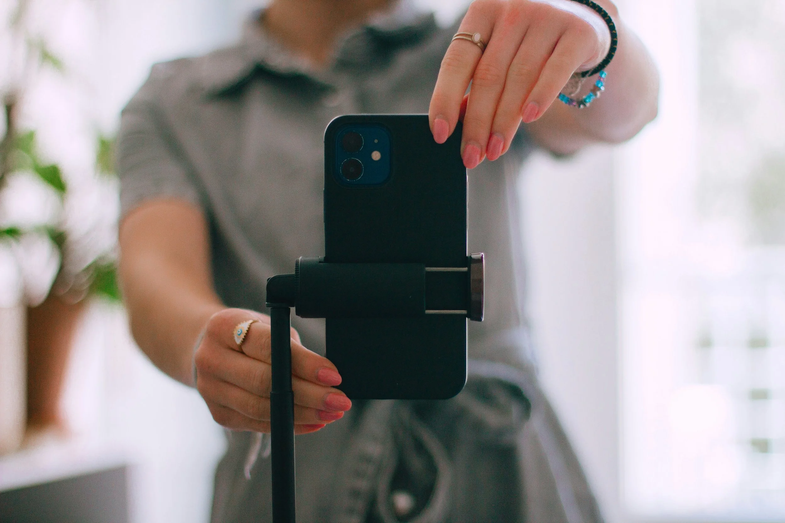 A person with painted nails is holding a smartphone in front of a mirror, taking a selfie. The person is wearing jewelry, including rings and bracelets. The background is blurred with natural light.