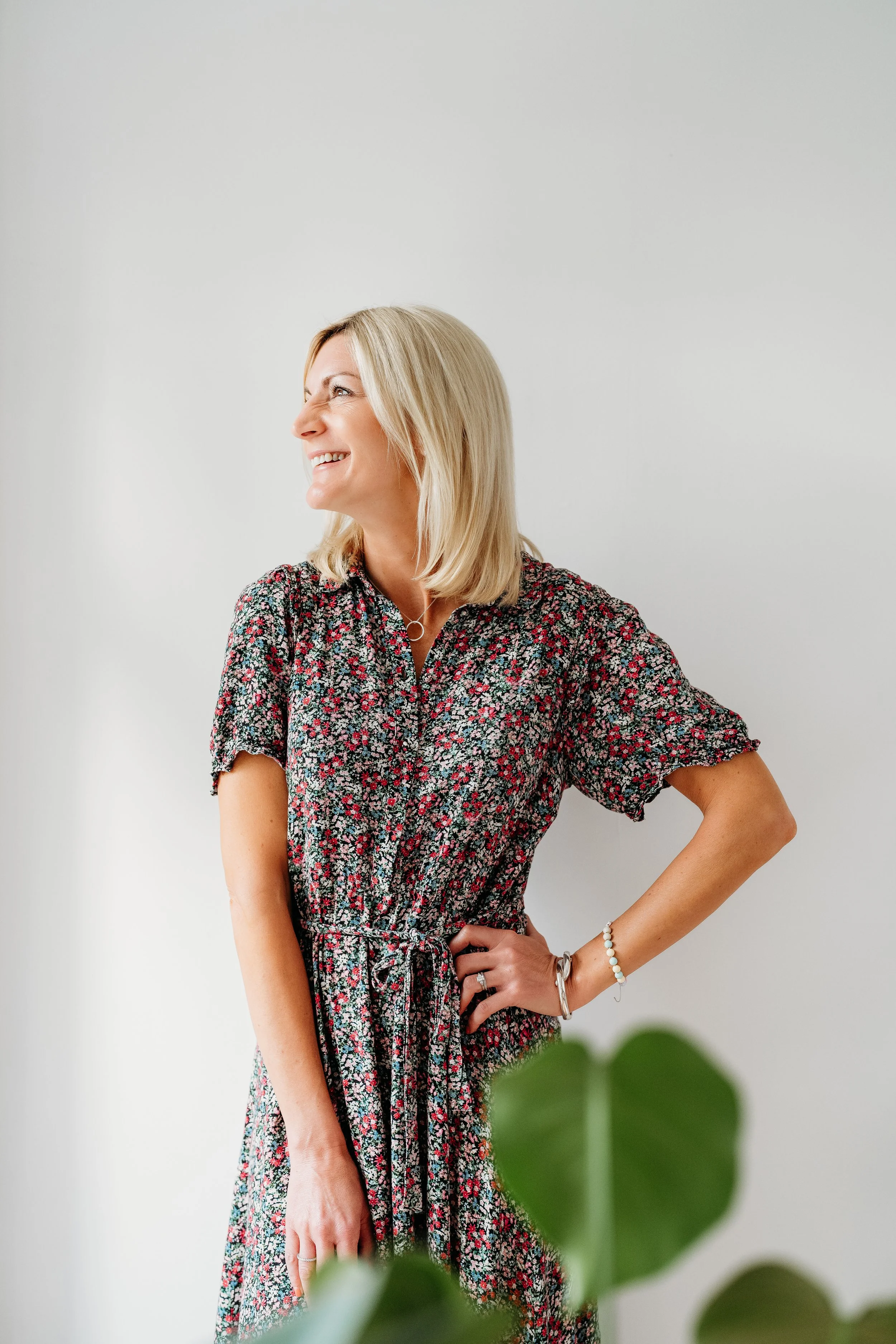A woman with shoulder-length blonde hair wearing a floral dress, smiling and looking to her left against a plain light-colored background.