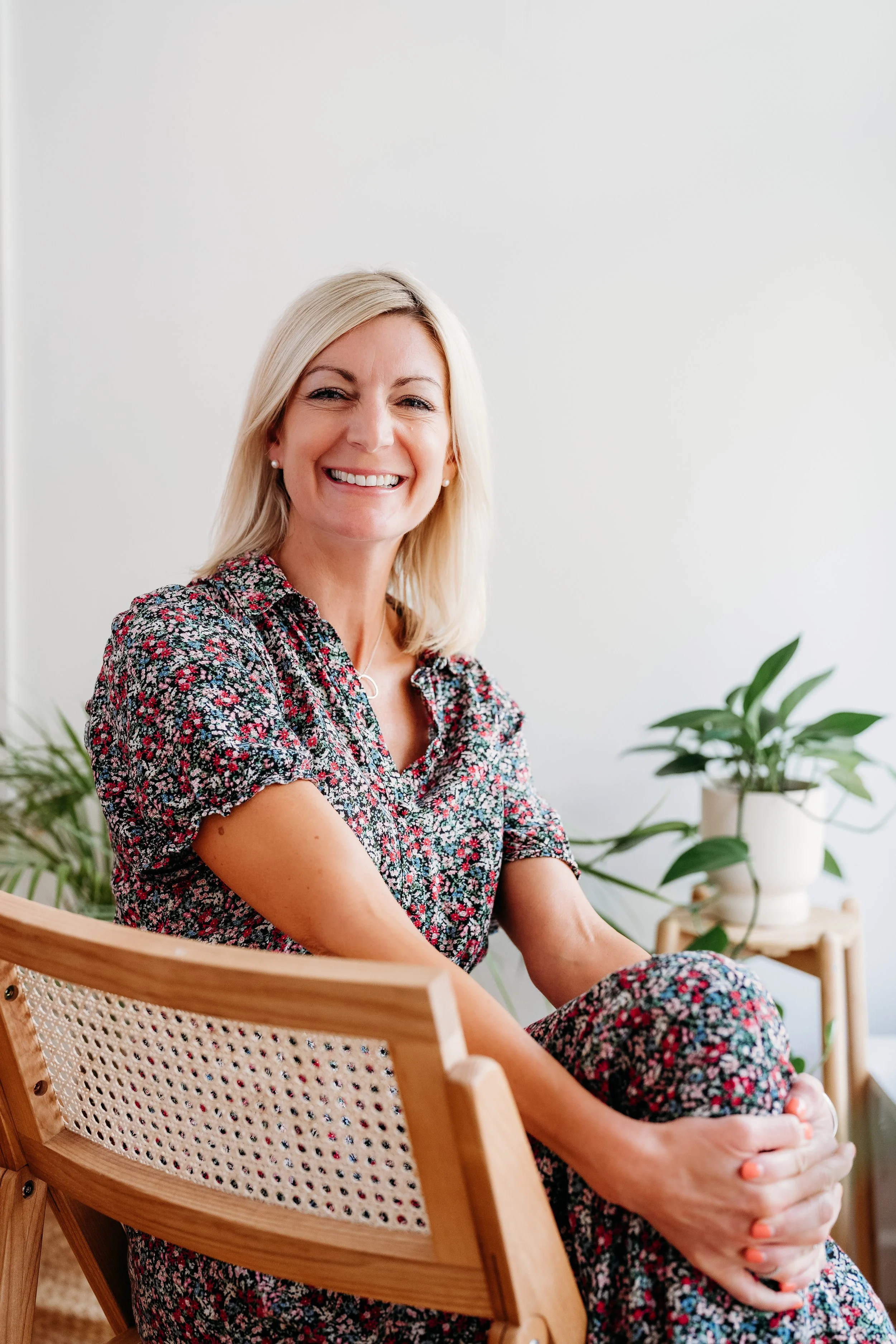 A woman with blonde hair, wearing a floral dress, sitting in a wicker chair, smiling with her eyes closed, with green plants in the background.