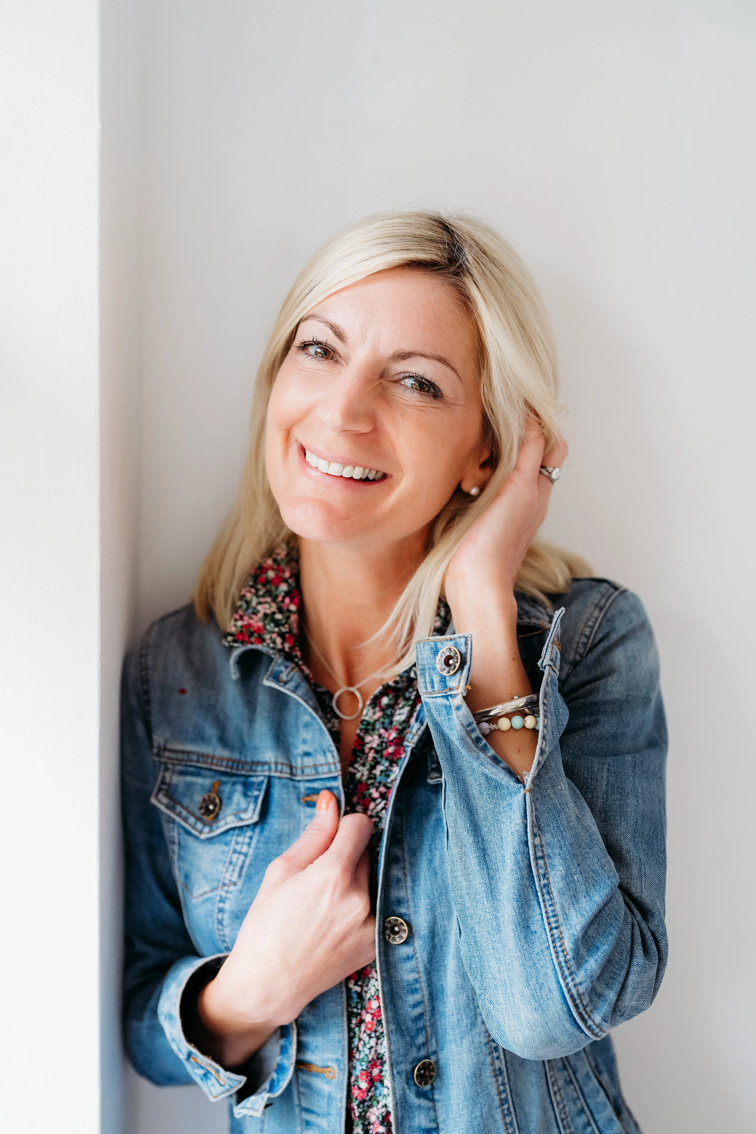 A woman smiling and looking at the camera, wearing a denim jacket over a floral shirt, with blonde hair and light skin, standing against a neutral background.