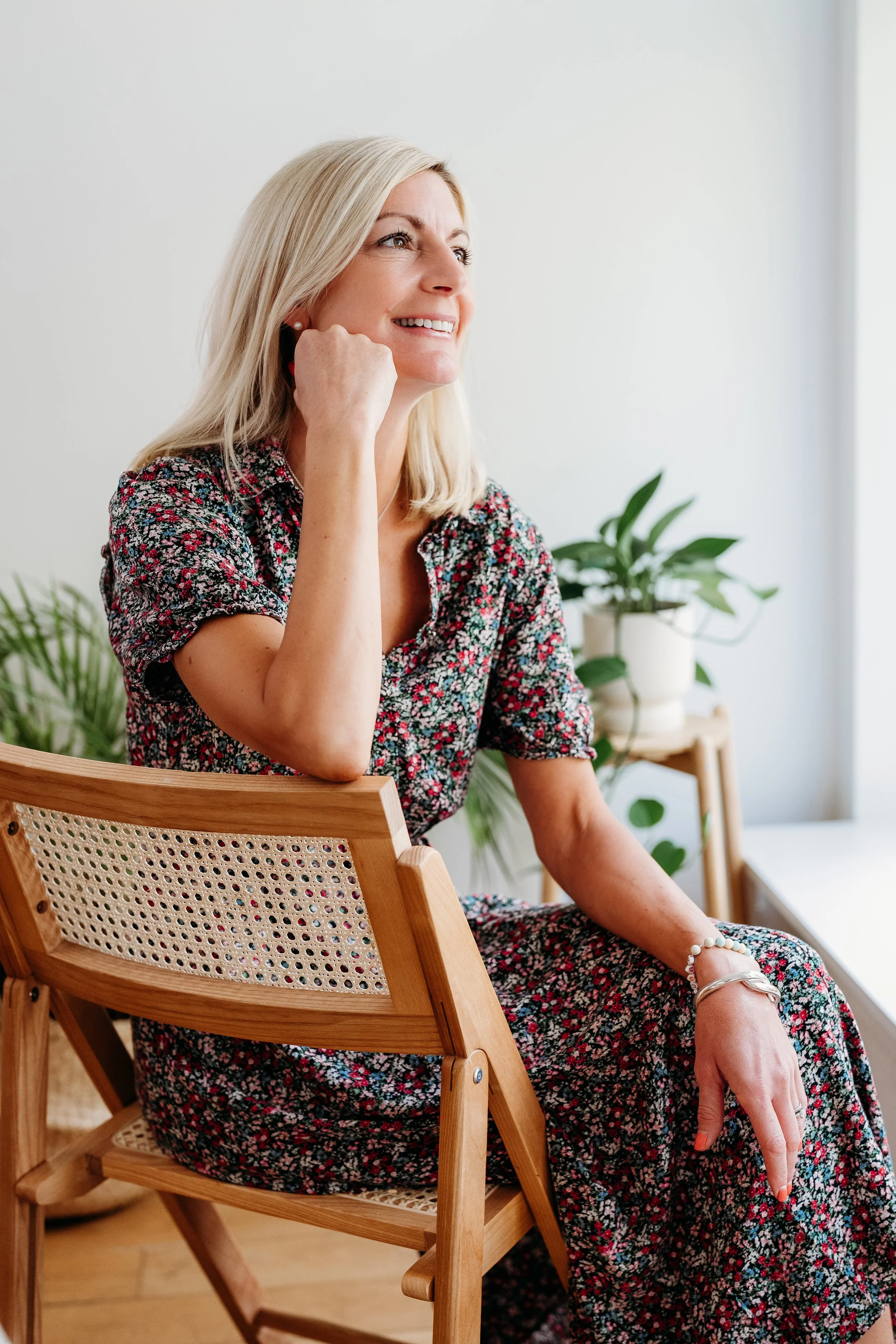 A middle-aged woman with blonde hair and a floral dress sitting on a wooden chair, smiling and looking slightly to her left, with potted plants in the background.