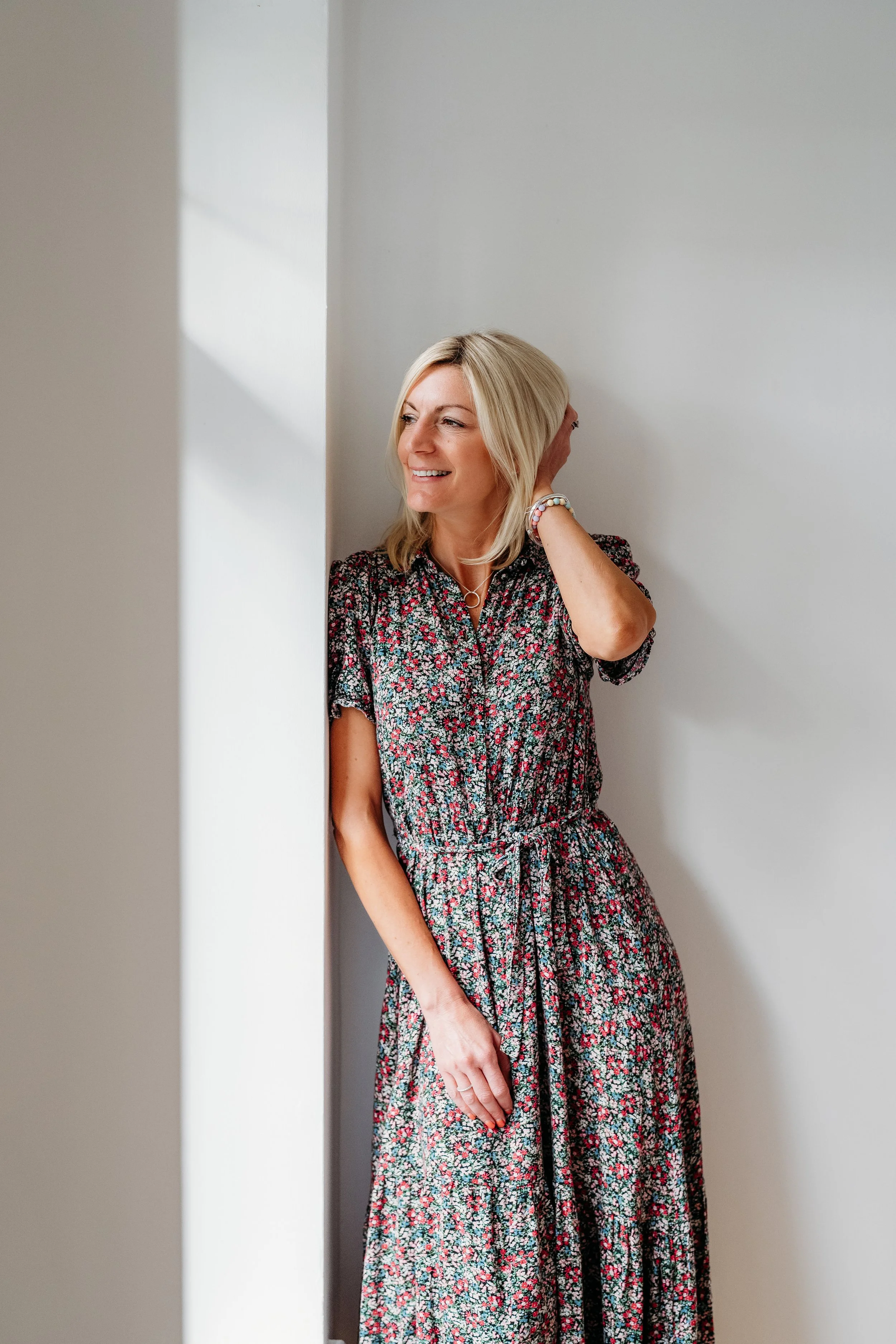 A woman with shoulder-length blonde hair smiling, wearing a floral dress and a beaded bracelet, standing with her hand behind her neck, leaning against a white wall in a well-lit room.