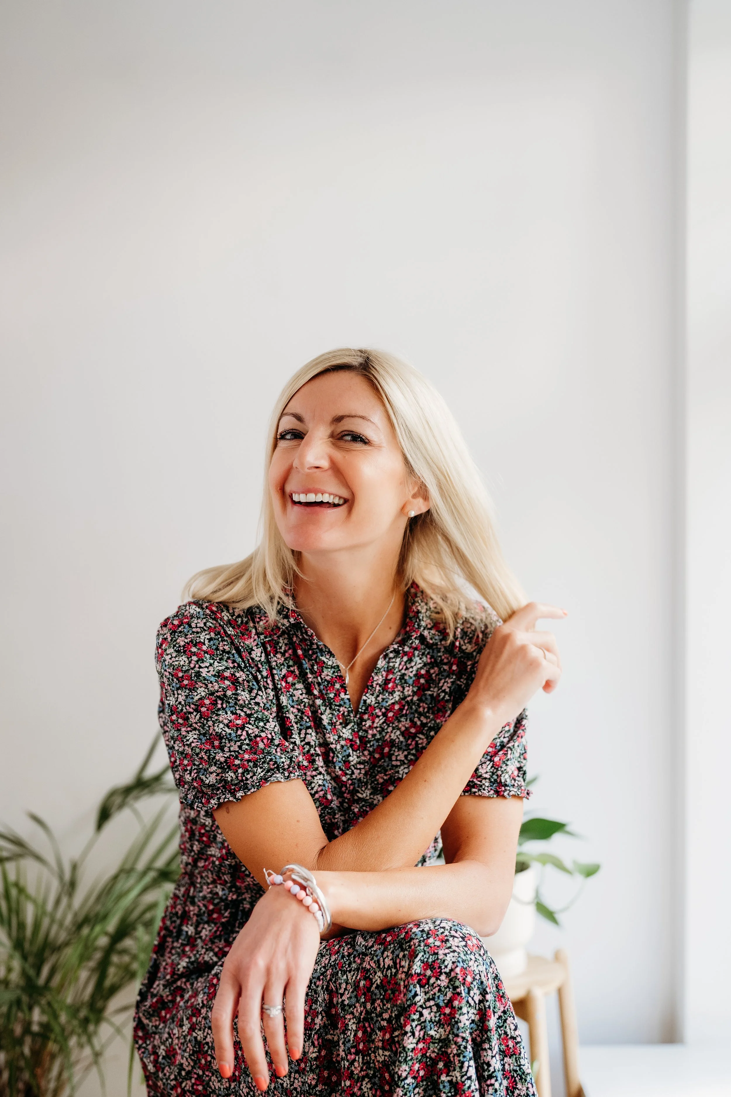 A smiling woman with blonde hair sitting in a bright room, wearing a floral dress, with a plant and a wooden stool in the background.