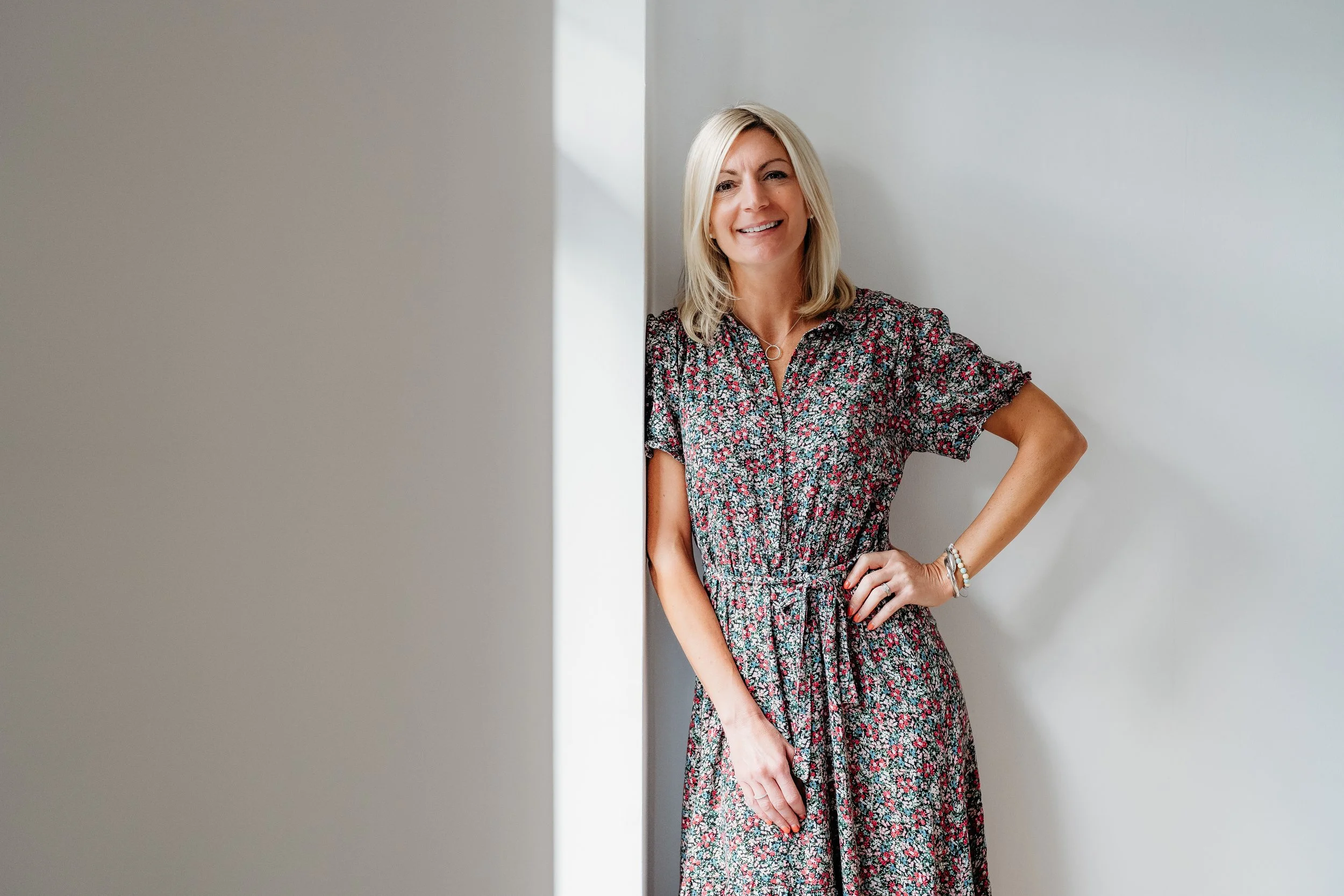 A smiling woman with blonde hair in a floral dress standing against a plain light-colored wall.