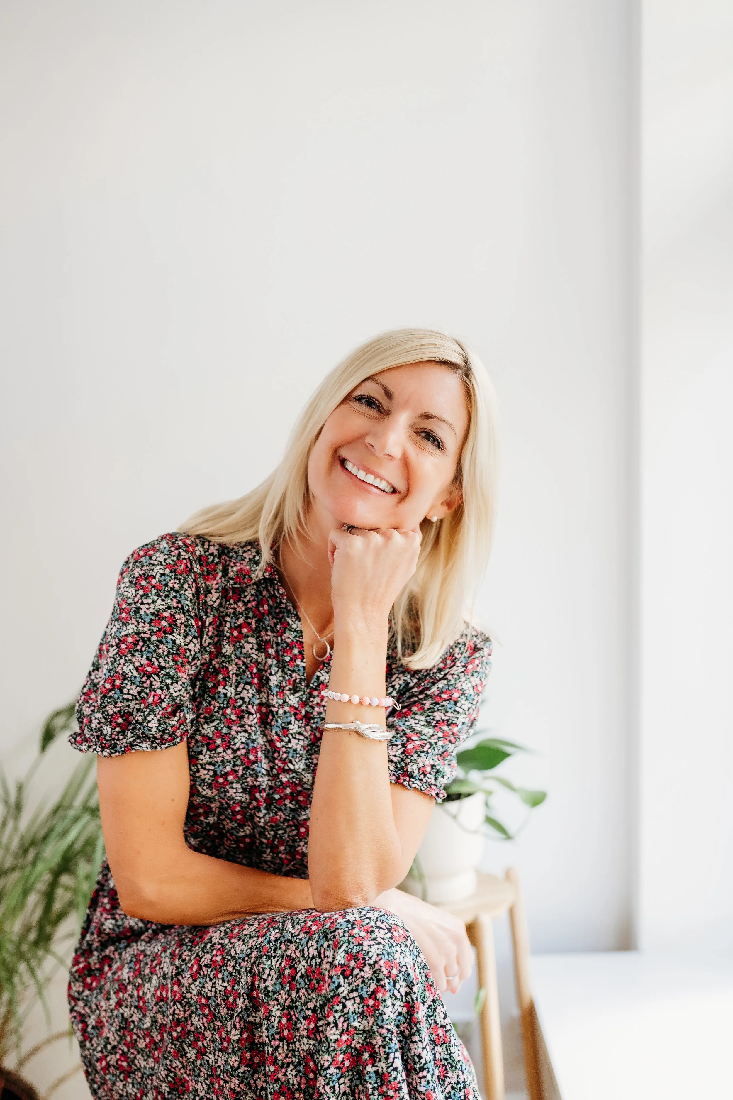 A smiling blonde woman wearing a floral dress, sitting with her elbow resting on her knee and her chin on her hand, in a well-lit room with a plant in the background.