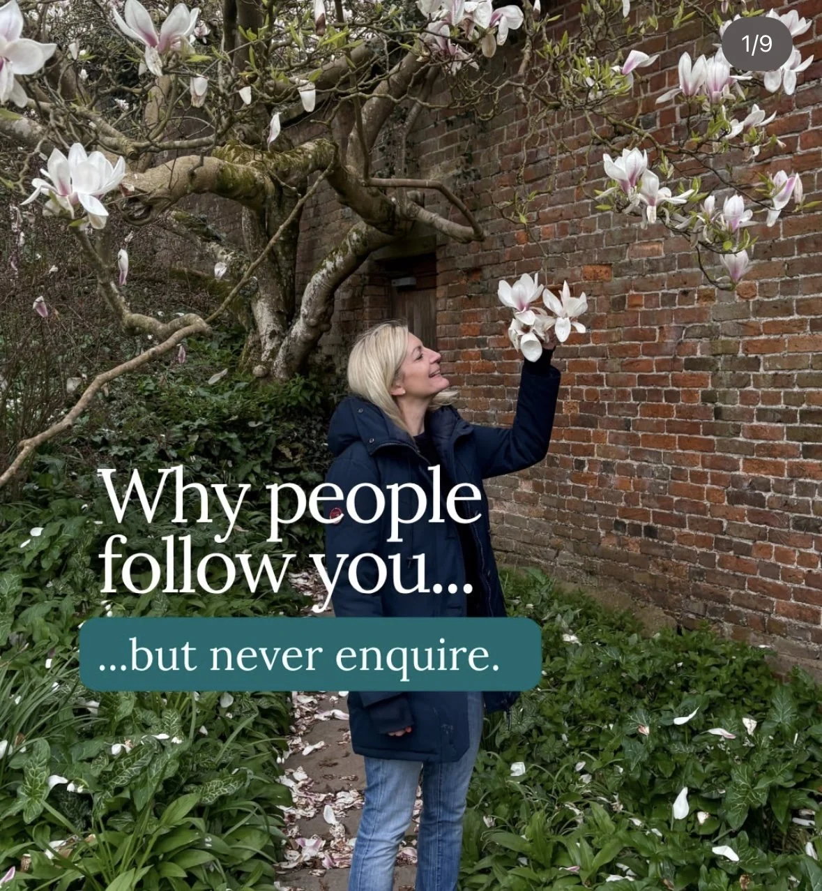 A woman with blonde hair wearing a dark jacket, standing outdoors next to a brick wall and flowering tree, smiling and reaching towards white and pink magnolia blossoms.