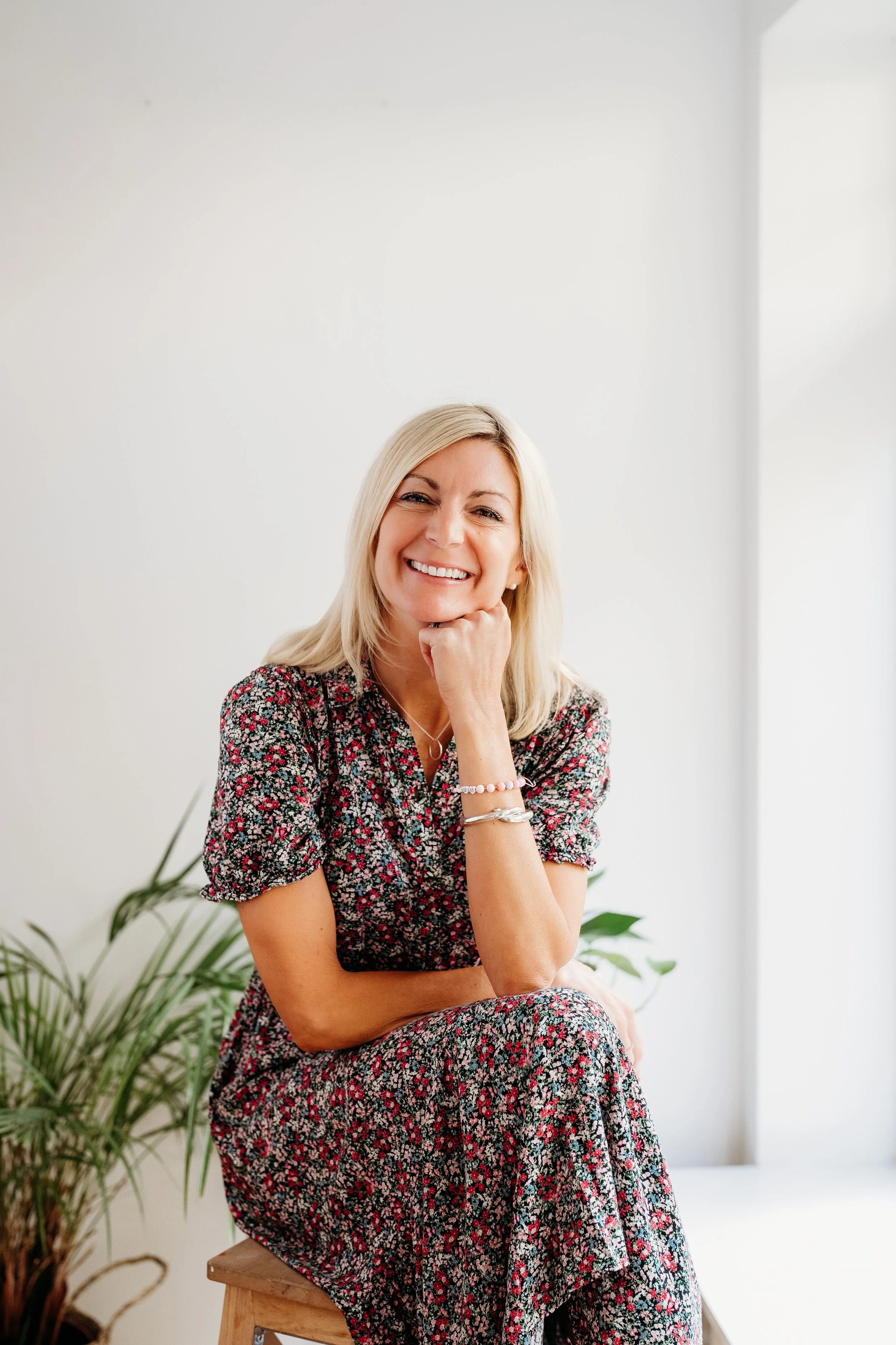 A woman with blonde hair smiling, wearing a floral dress, sitting on a wooden stool with one hand resting on her chin, in a bright room with white walls and green plants in the background.