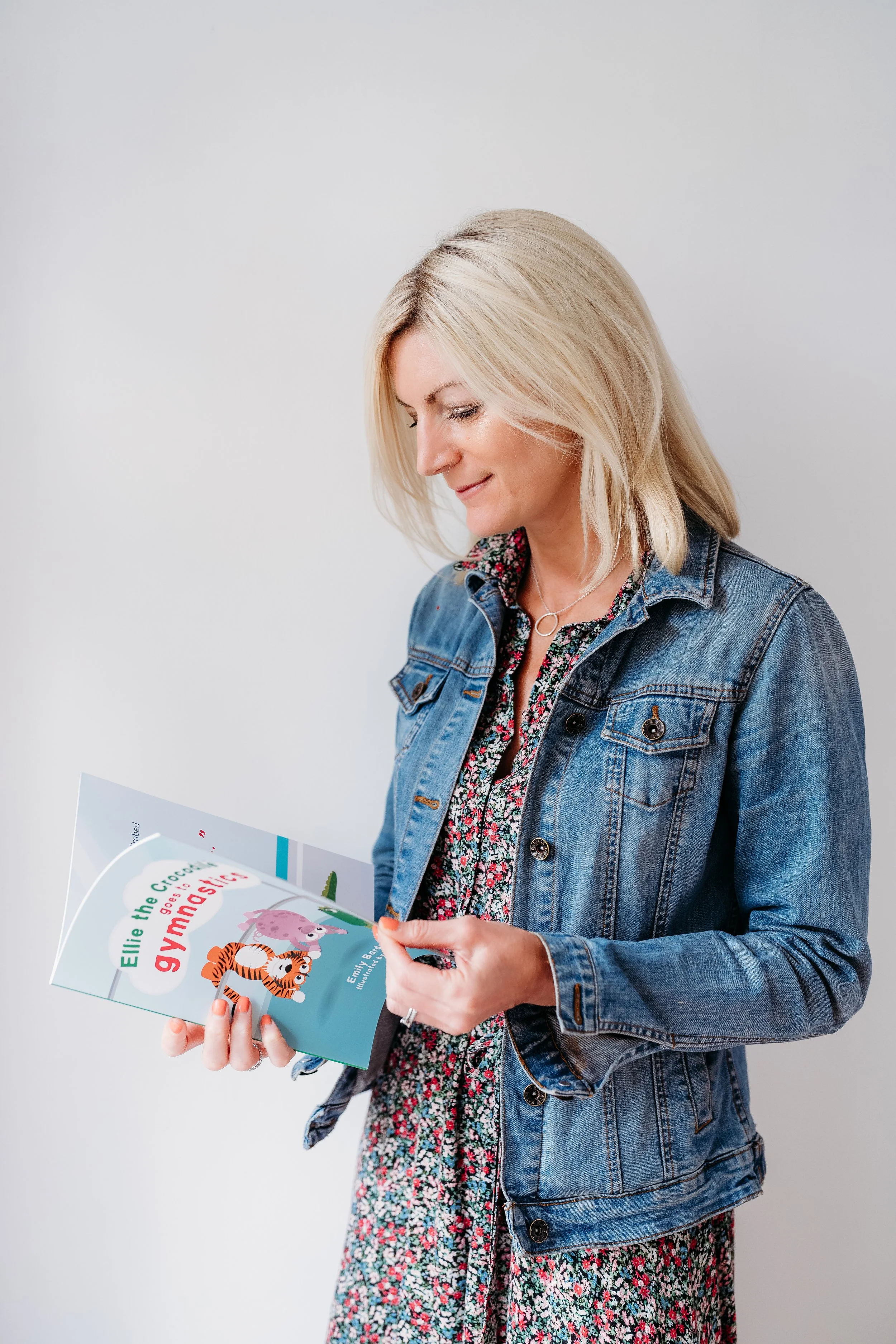 A woman with blonde hair wearing a denim jacket looking at a children's book titled 'Elle the Crocodile goes to gymnastics' with a cartoon tiger and elephant on the cover.