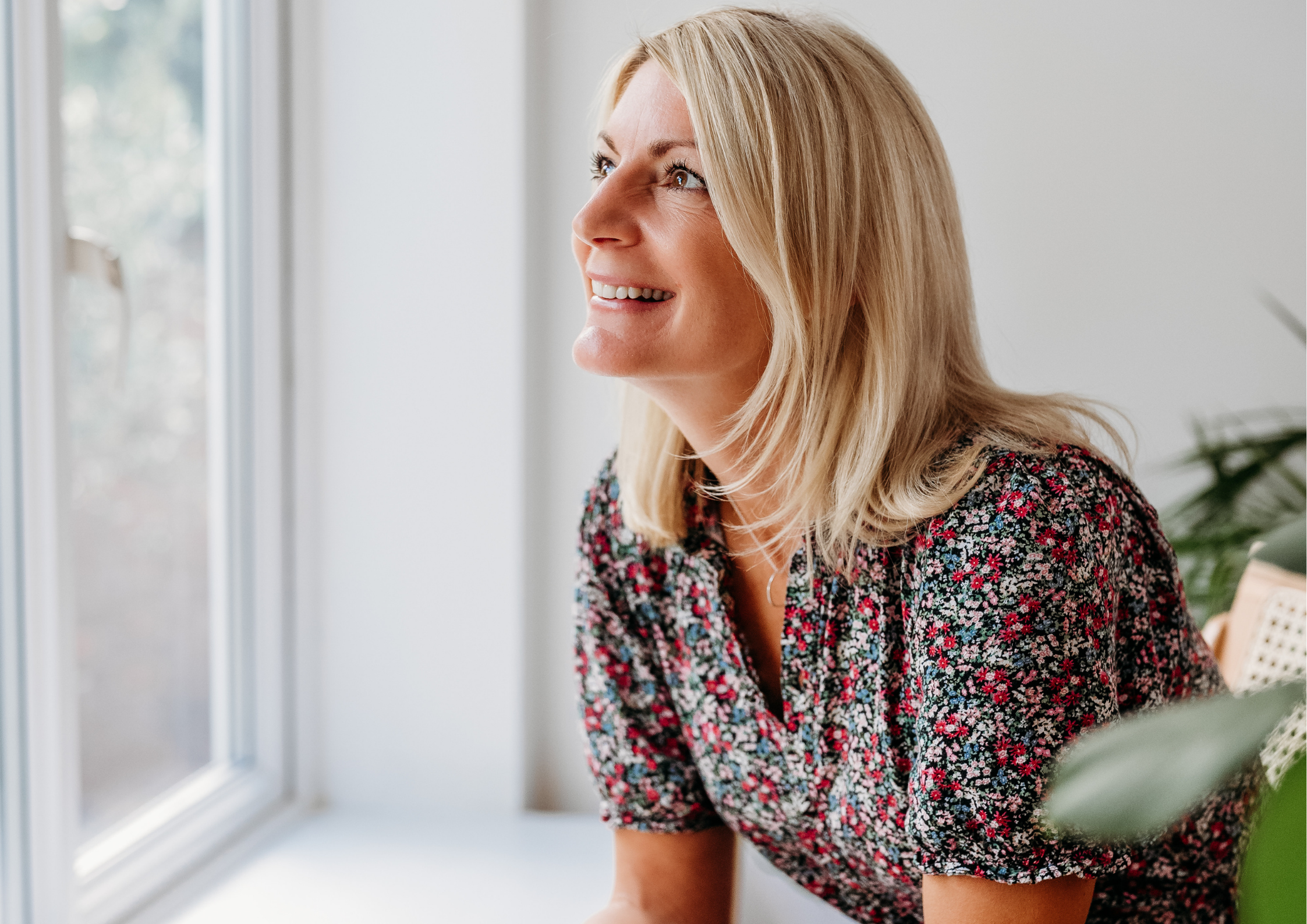 A smiling woman with blonde hair wearing a floral dress, looking out of a window in a brightly lit room.