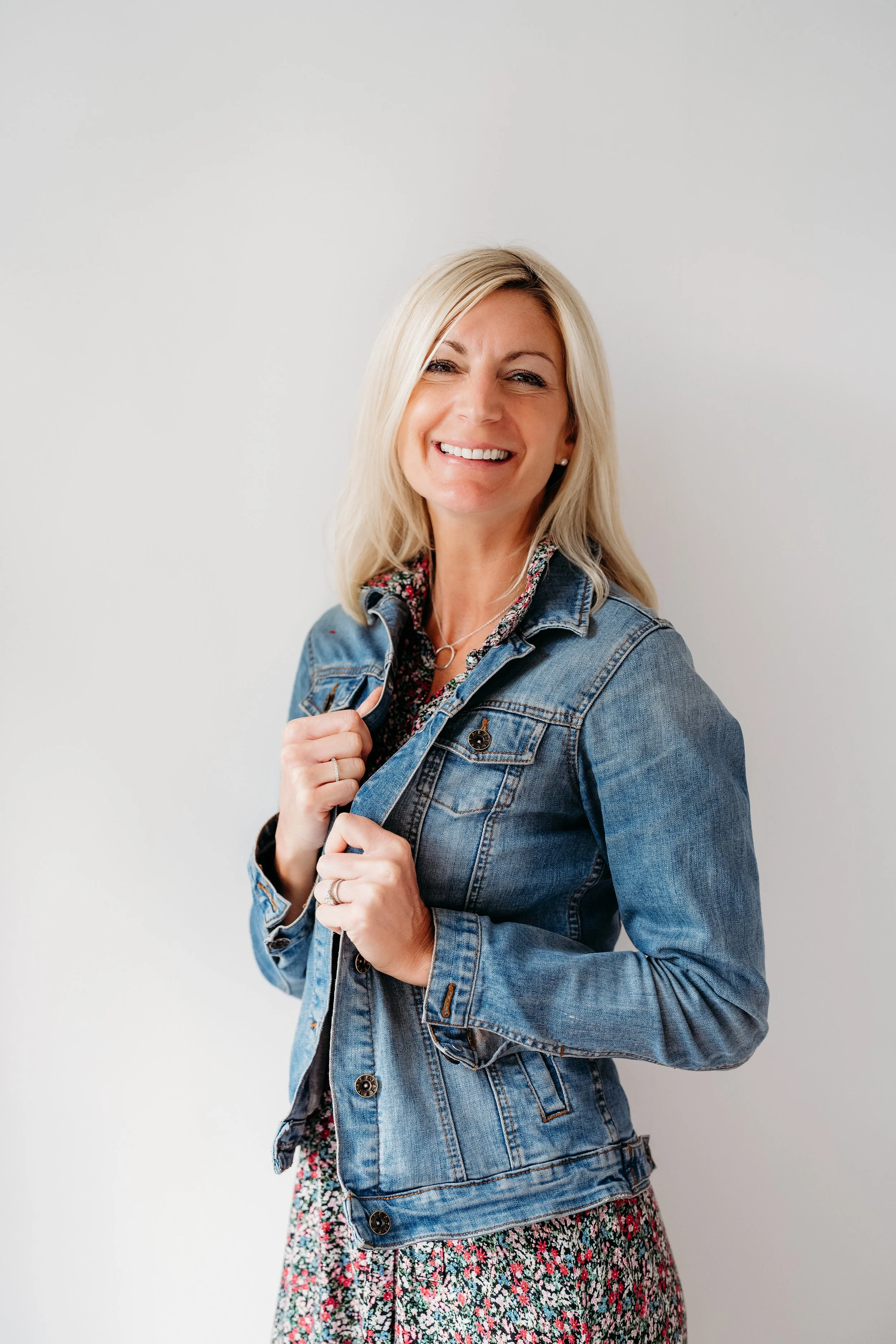 A woman with blonde hair smiling, wearing a denim jacket over a floral dress, standing against a plain white background.