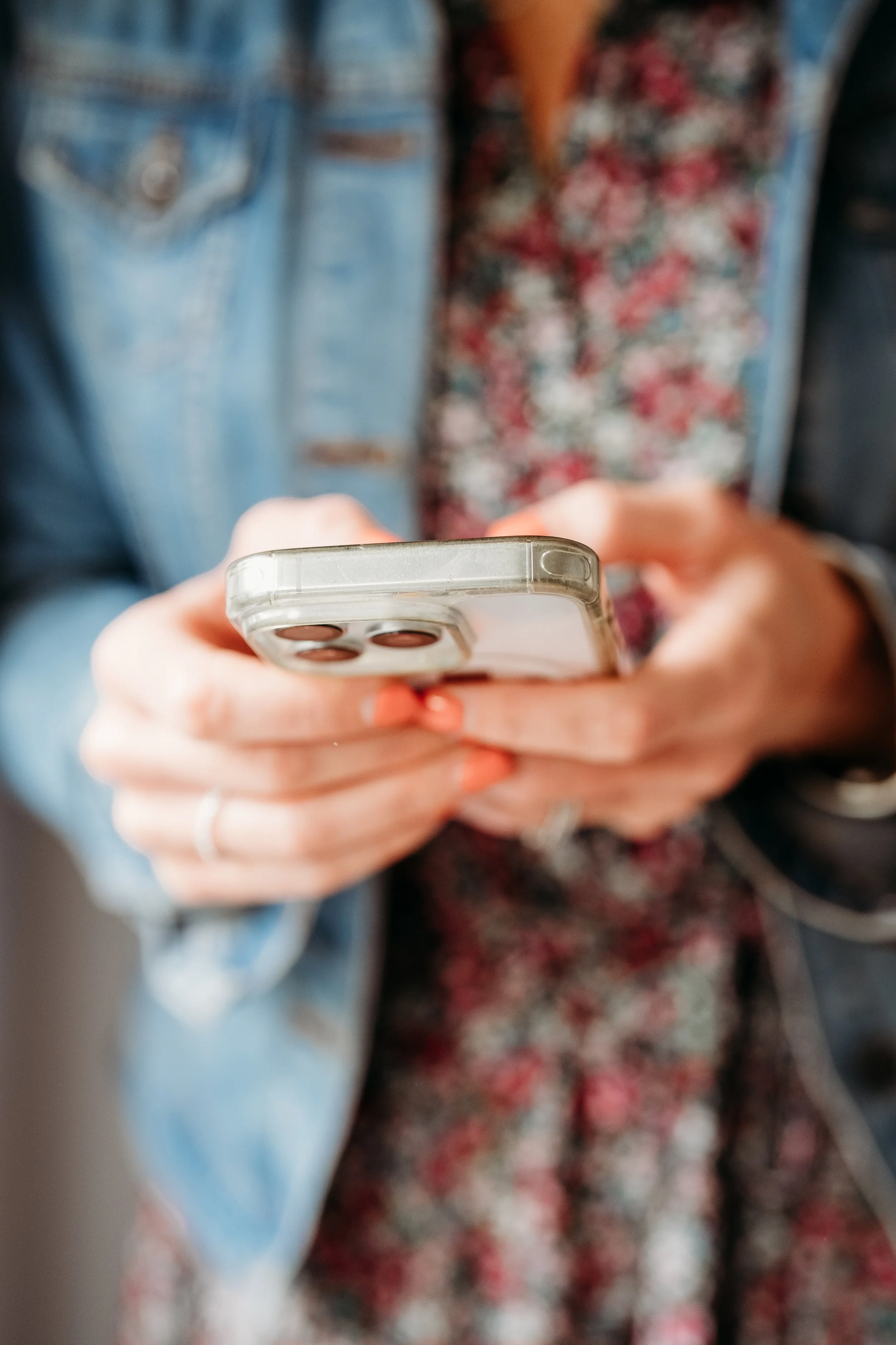 Close-up of a person's hands holding a smartphone with a quad-camera setup, blurred background showing denim and floral clothing.