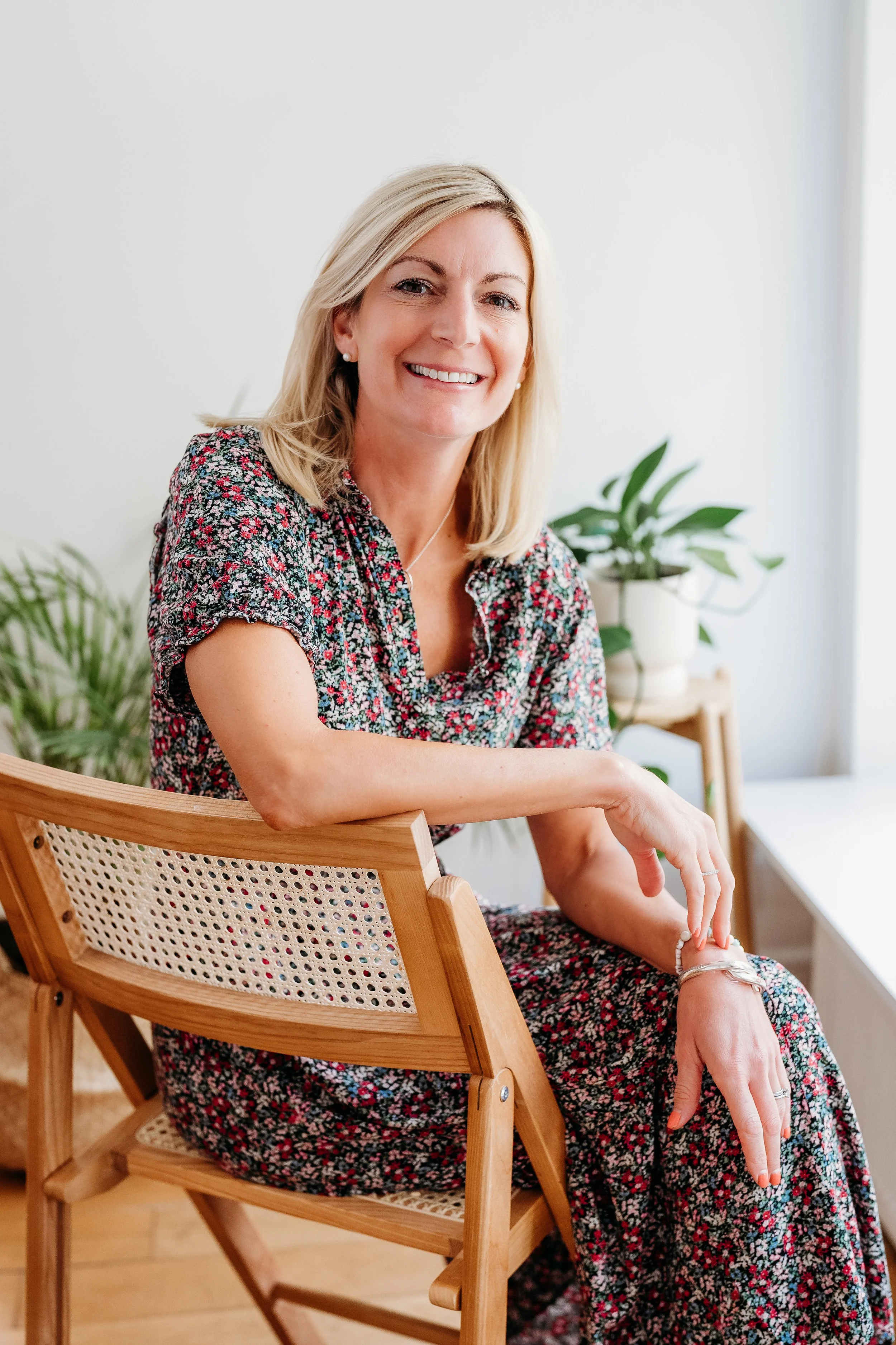 A smiling woman with blonde hair, wearing a floral dress, sitting on a wooden chair in a bright room with green plants in the background.