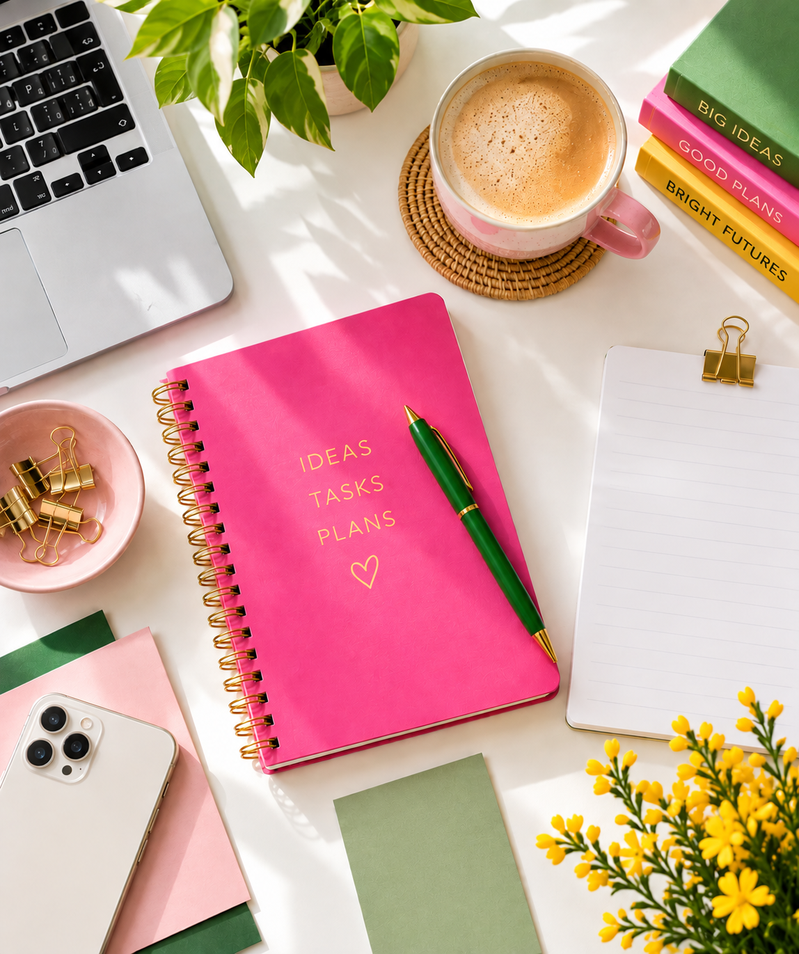 A desk workspace with a pink planner labeled 'Ideas Tasks Plans', a green pen, a white notebook, a cup of coffee, a plant, a laptop, a smartphone, colorful books, gold binder clips, and yellow flowers.