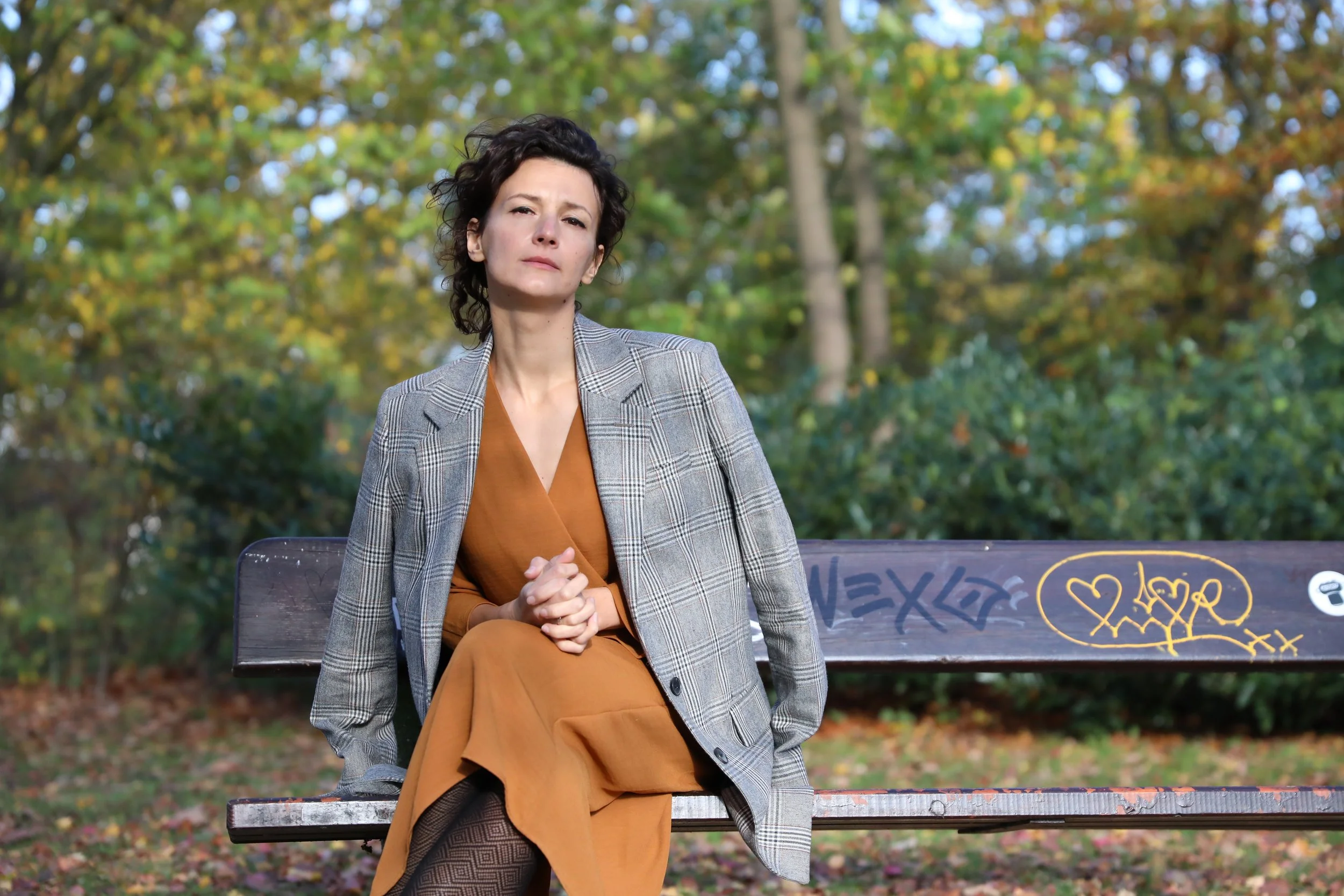 A woman with short dark hair, wearing a gray checkered blazer and a brown dress, sitting on a graffiti-covered park bench in an outdoor setting with trees and fall foliage in the background.