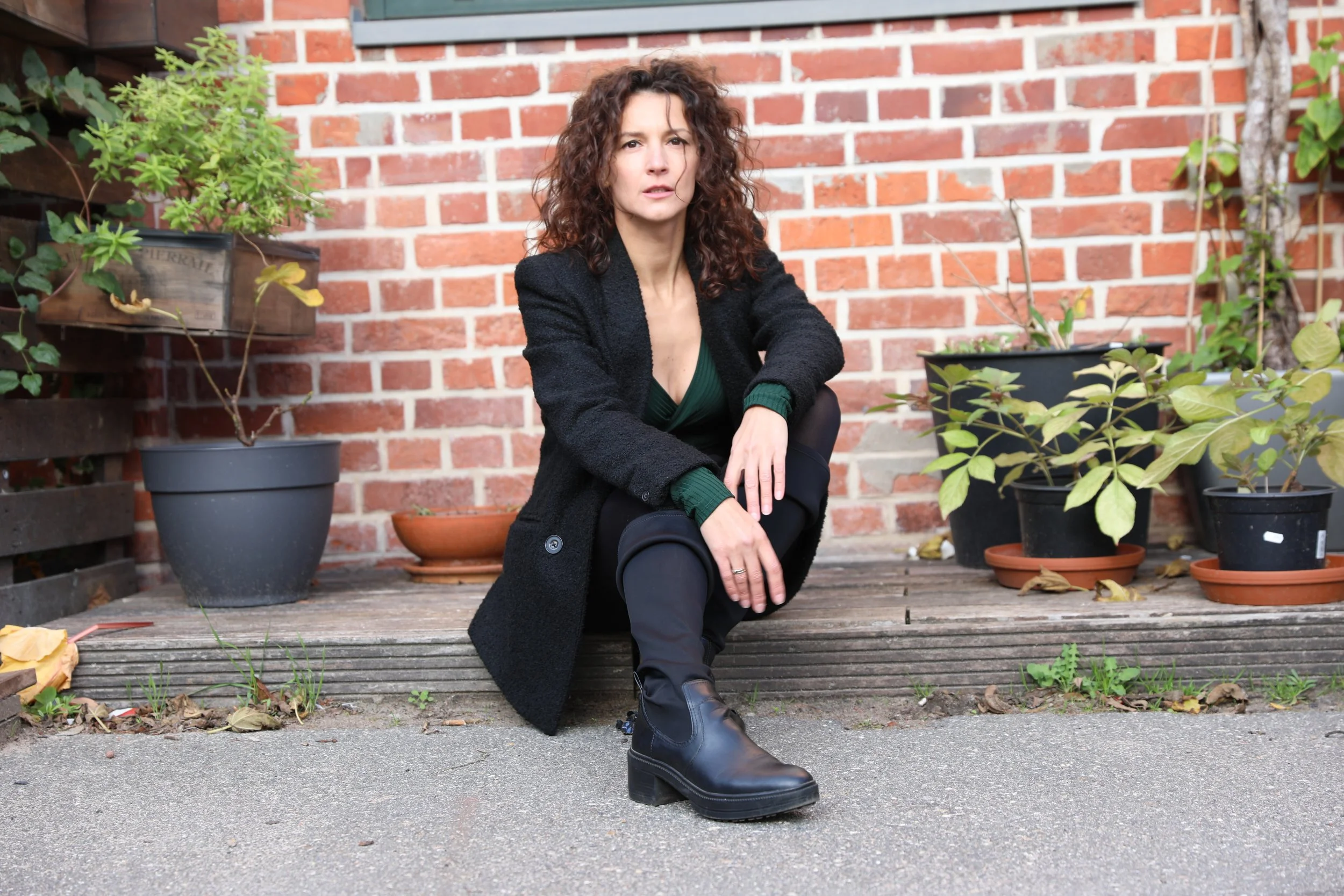 A woman with curly brown hair sitting on a wooden platform in front of a brick wall, wearing a black coat, green sweater, black pants, and black boots, surrounded by potted plants.