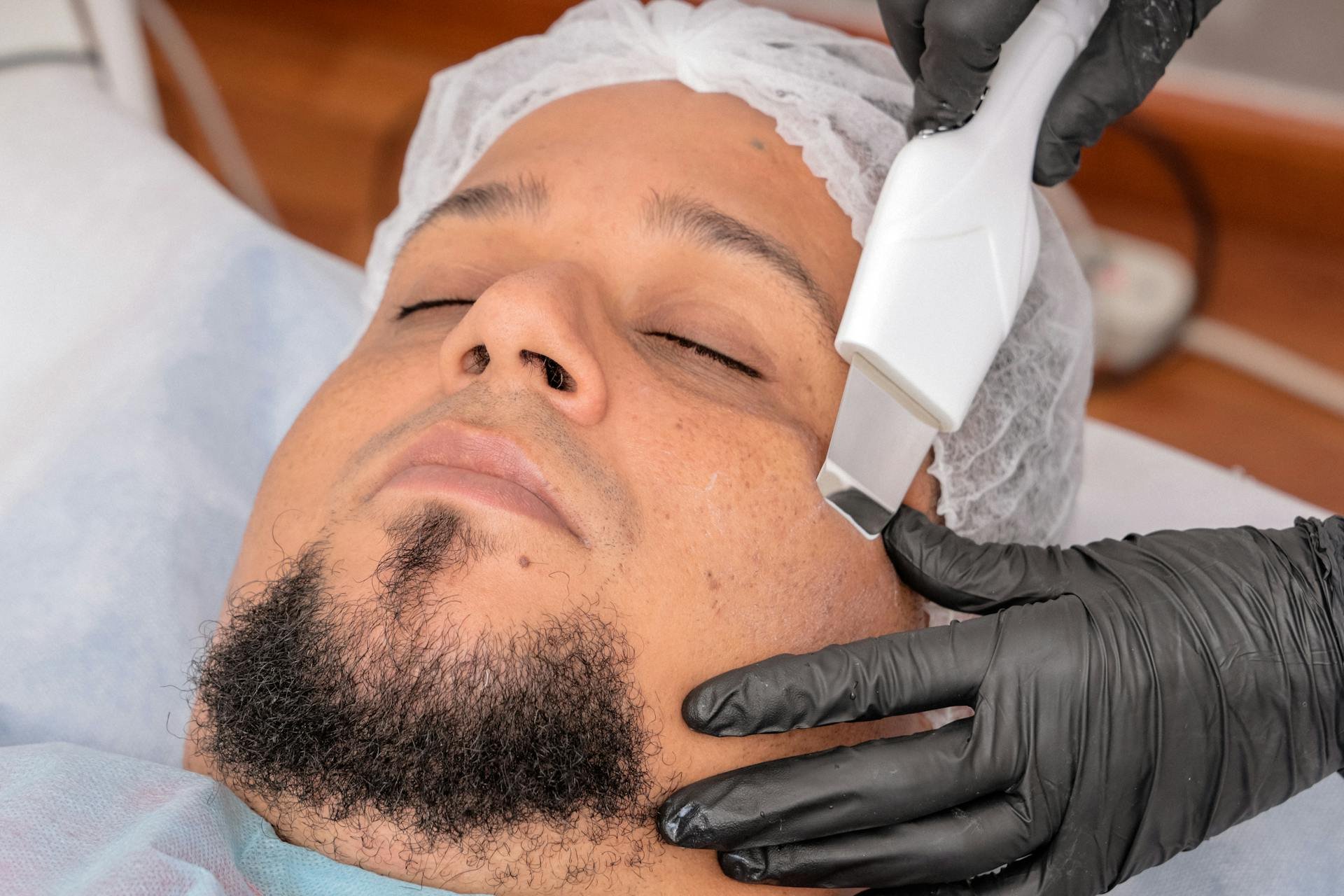 A man lying down with eyes closed, receiving a facial treatment from a healthcare professional wearing black gloves and using a handheld device on his face.