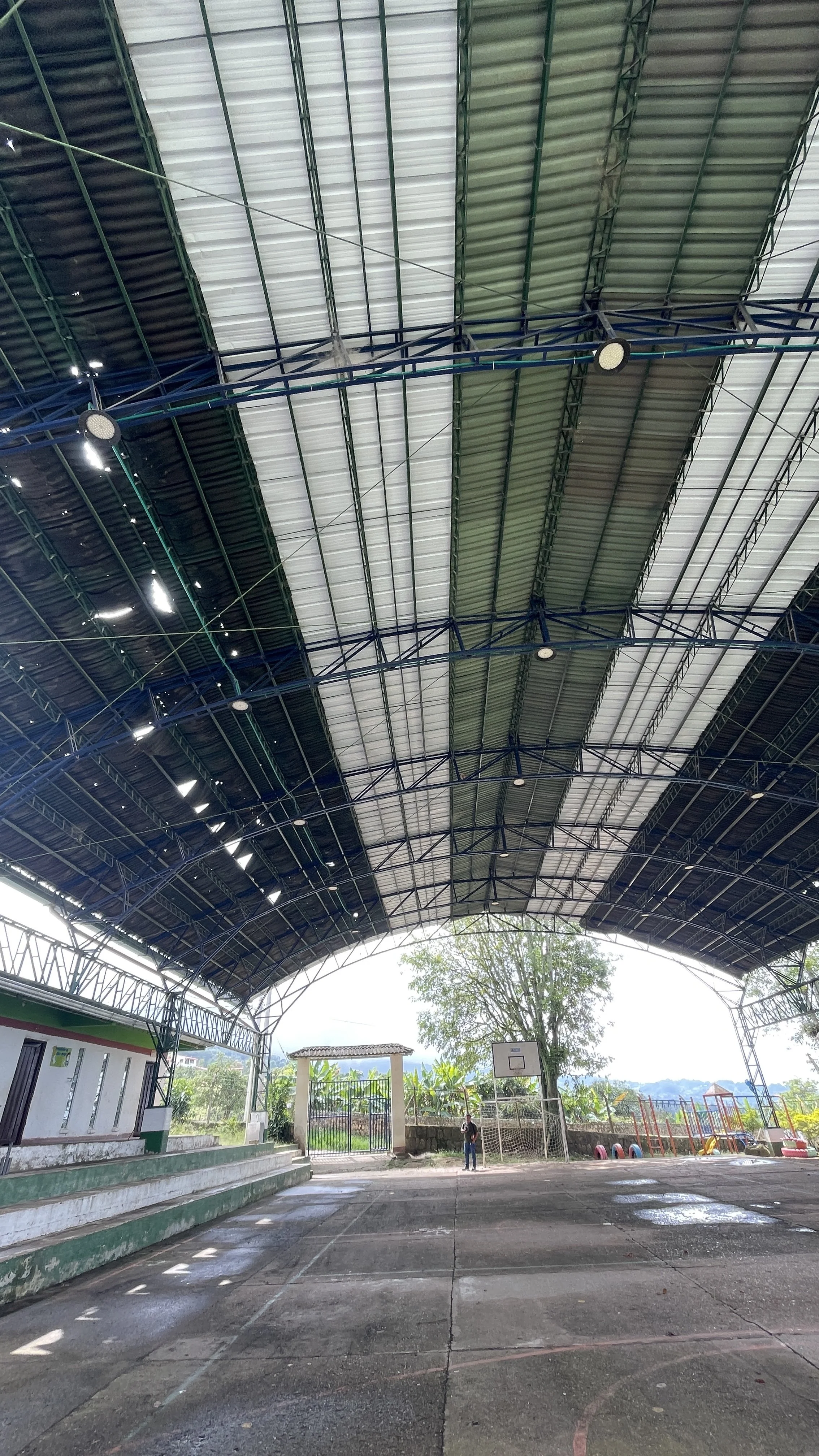 Here the school facilities. Roof damage. An indoor basketball court with a metal roof, a basketball hoop, and playground equipment in the background.