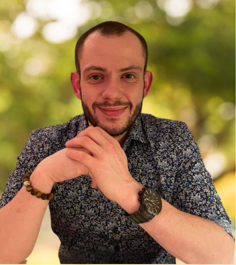 A smiling man with short hair, a beard, and a floral shirt, sitting outdoors with a blurred green background. David, the treasurer of the assosiation.