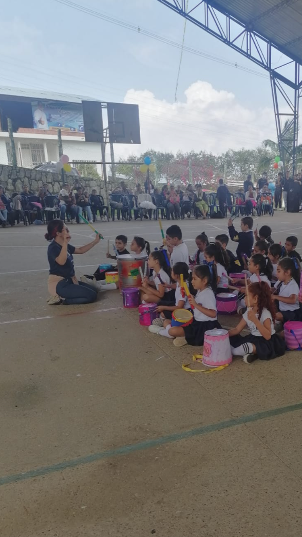 Children sitting on the ground playing drums with a woman instructor during a school event or performance at a covered outdoor sports court.
