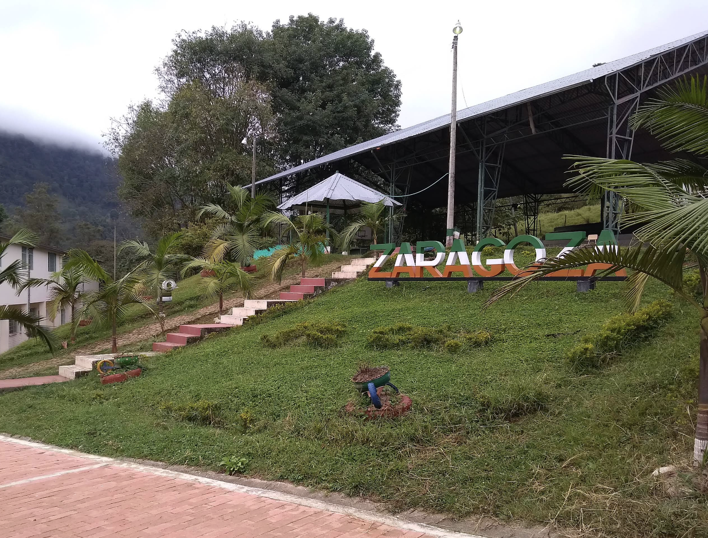 Outdoor stage with a large sign reading 'ZARAGOZA' on a grassy hill, surrounded by small palm trees, with a misty mountain in the background.