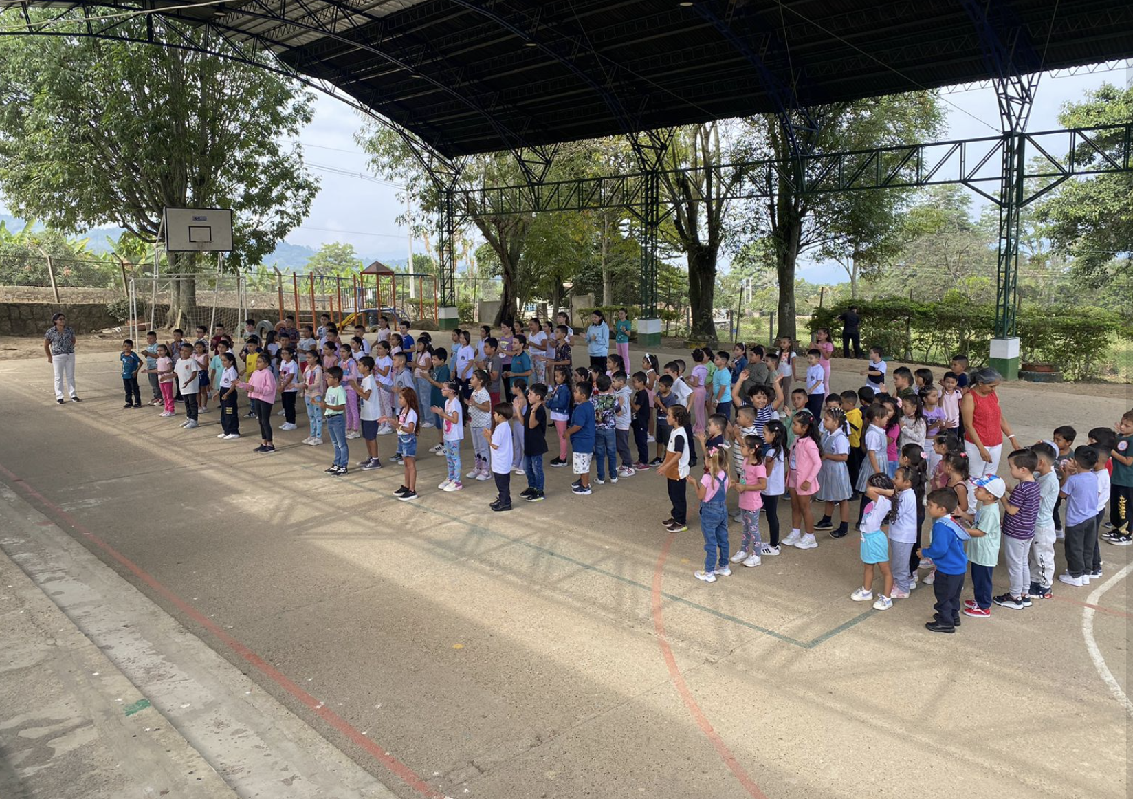 A large group of children gathered in an indoor basketball court, standing in organized rows under a high roof, with a few adults supervising.  This are children supported by seeds for a better world asbl.