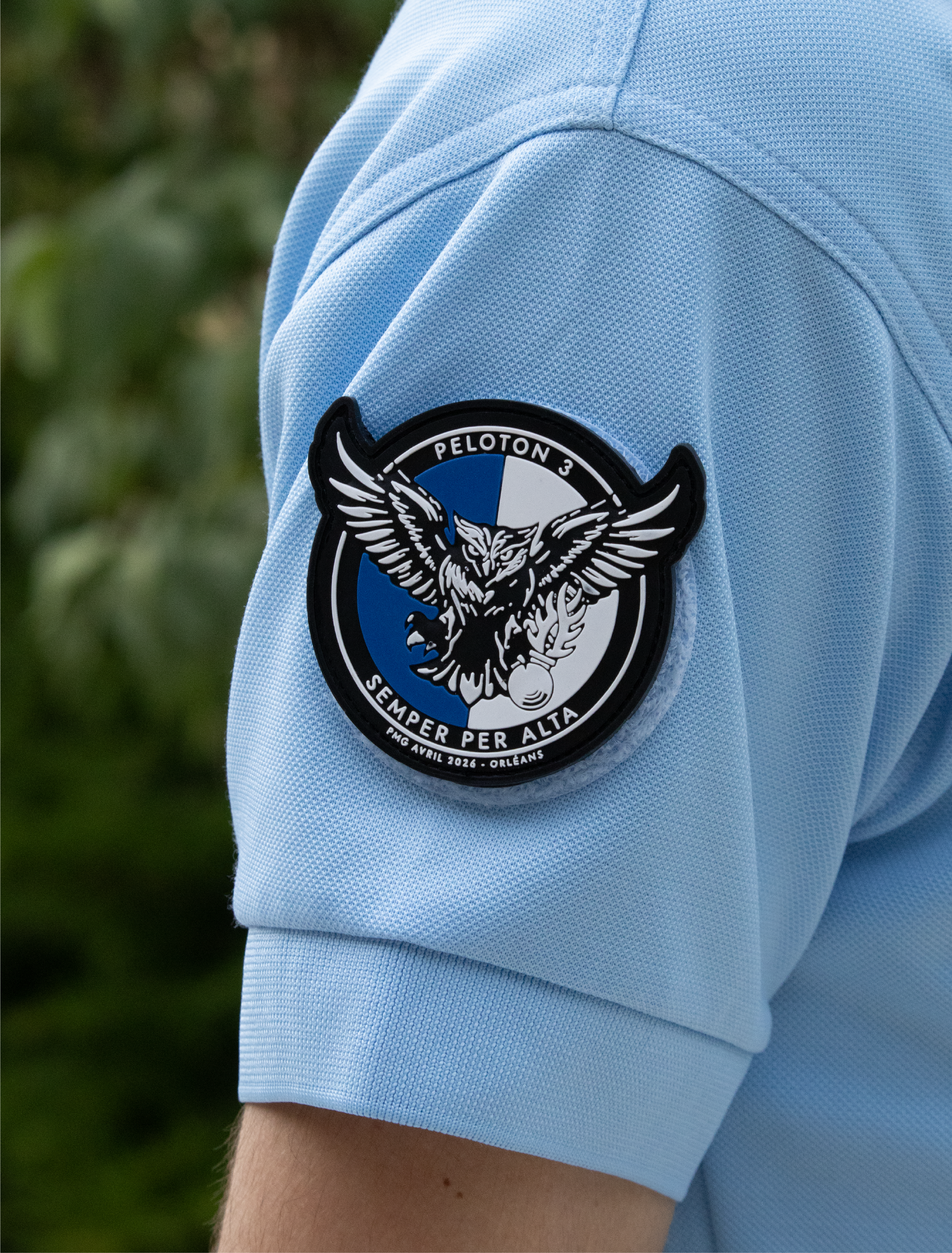 Le bras d'une personne portant un maillot de sport bleu clair avec un écusson de l'équipe de football à l'épaule, dans un environnement extérieur