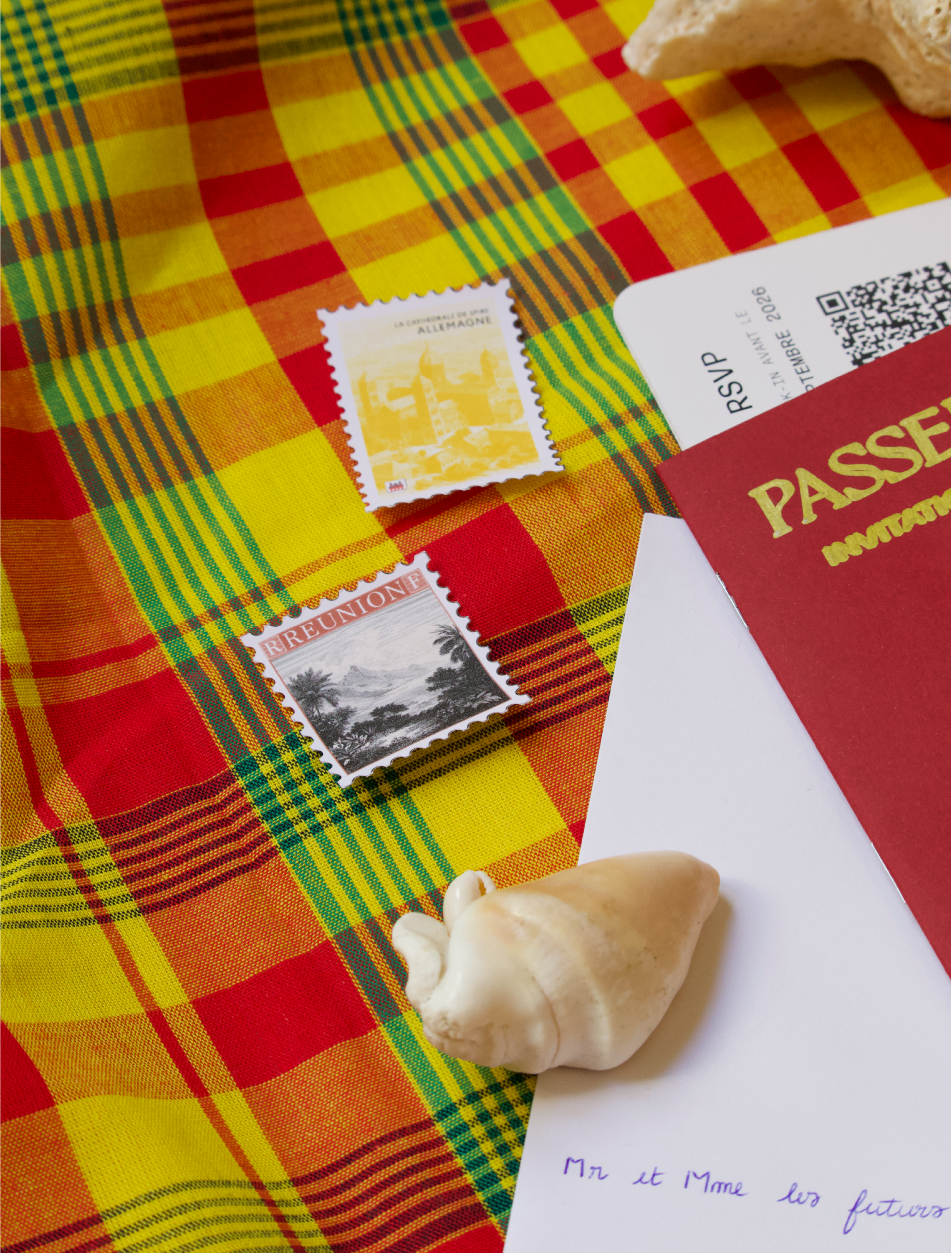 Deux timbres postaux, un avec une scène en noir et blanc de paysages avec des arbres, et un autre en jaune avec une scène de ville d'Allemagne, sont posés sur un tissu à carreaux rouge, jaune, vert et noir. Une partie d'un passeport rouge est visible à droite avec le texte 'PASS', ainsi qu'une note écrite à la main en français.