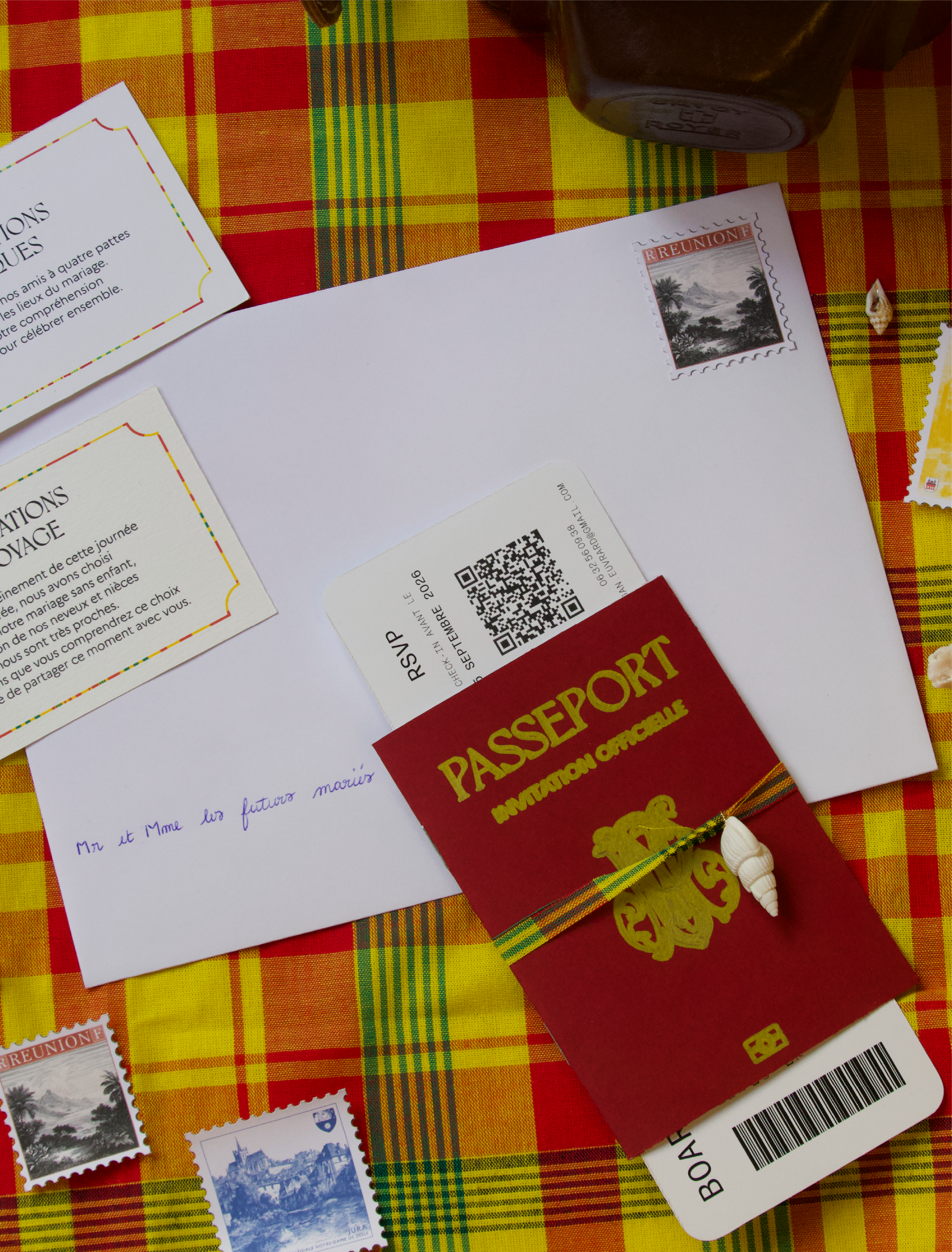 Une scène de bureau avec une enveloppe, un billet de voyage, des timbres, un téléphone portable, des papiers, et un tissu à carreaux rouge-jaune-vert.