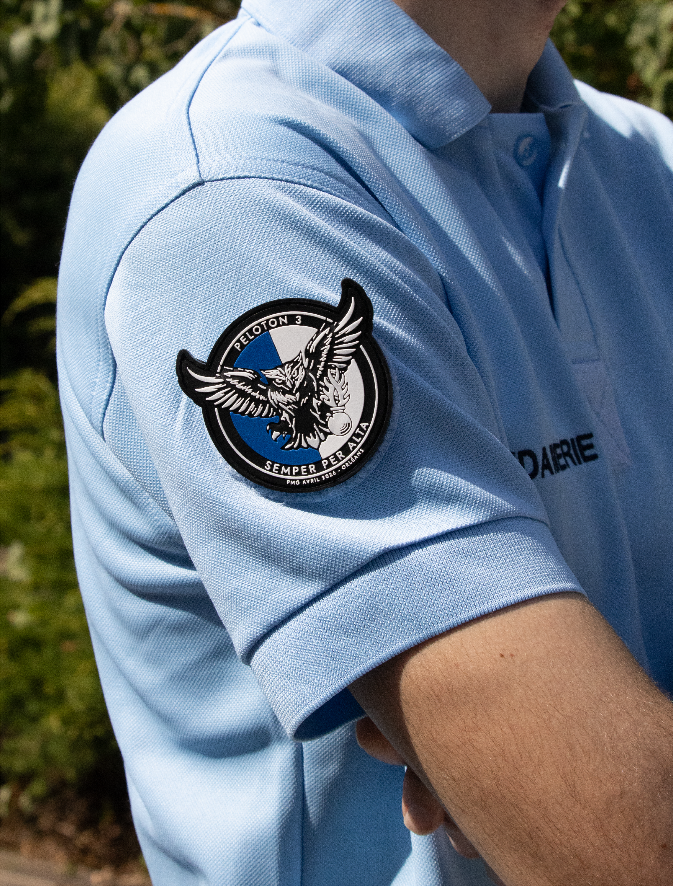 Close-up de l'épaule d'un agent de sécurité portant un uniforme bleu clair avec un patch rond sur l'épaule. Le patch montre un hibou avec des ailes déployées et un message en Latin. La personne est en extérieur avec un fond de végétation verte.