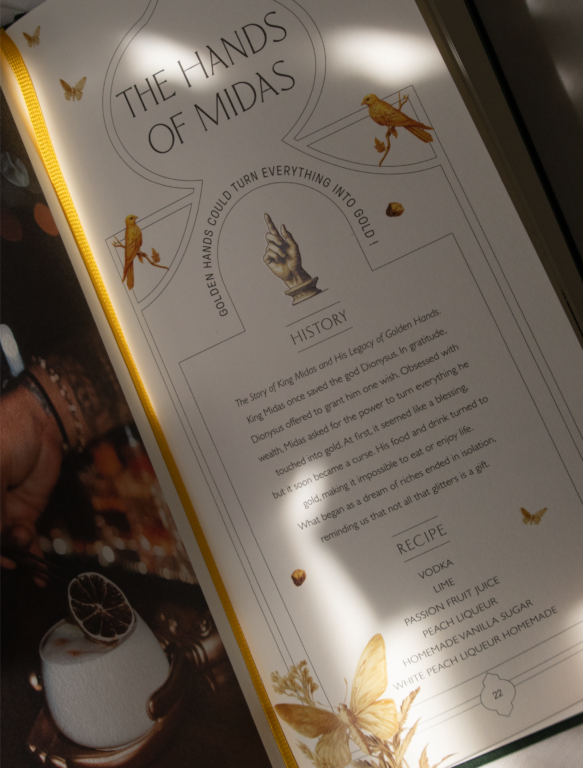 Une page d'un menu ou d'une brochure avec un titre 'The Hands of Midas' et un sous-titre 'Golden Hands Could Turn Everything Into Gold !' accompagnée d'illustrations d'oiseaux, de papillons et de faune en teintes dorées, avec une section sur l'histoire et une autre sur des recettes de cocktails.