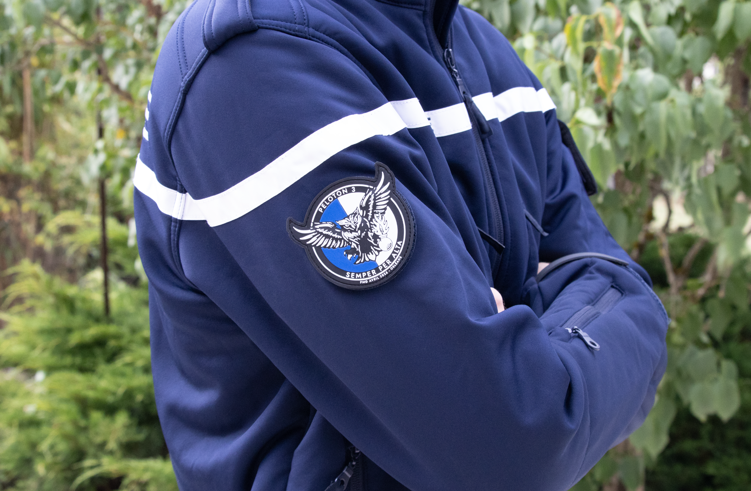 Un uniforme bleu marine avec une bande blanche porte un badge d'escouade de police ou de sécurité, avec un logo d'aigle et la phrase "Peloton 3 Semper Per Alia".